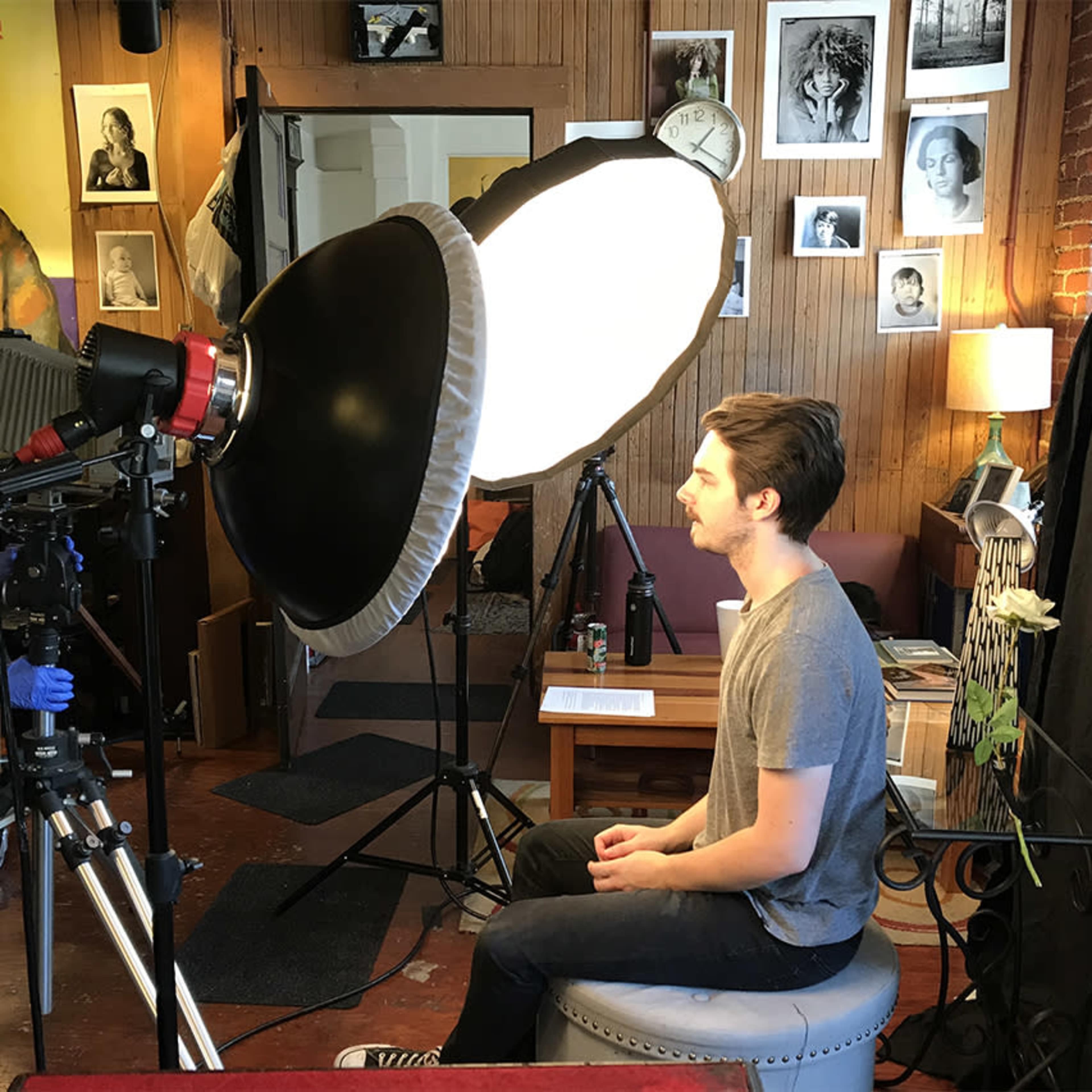 A photographer adjusts a large light source while a seated subject poses in a studio filled with various decor and framed photographs.