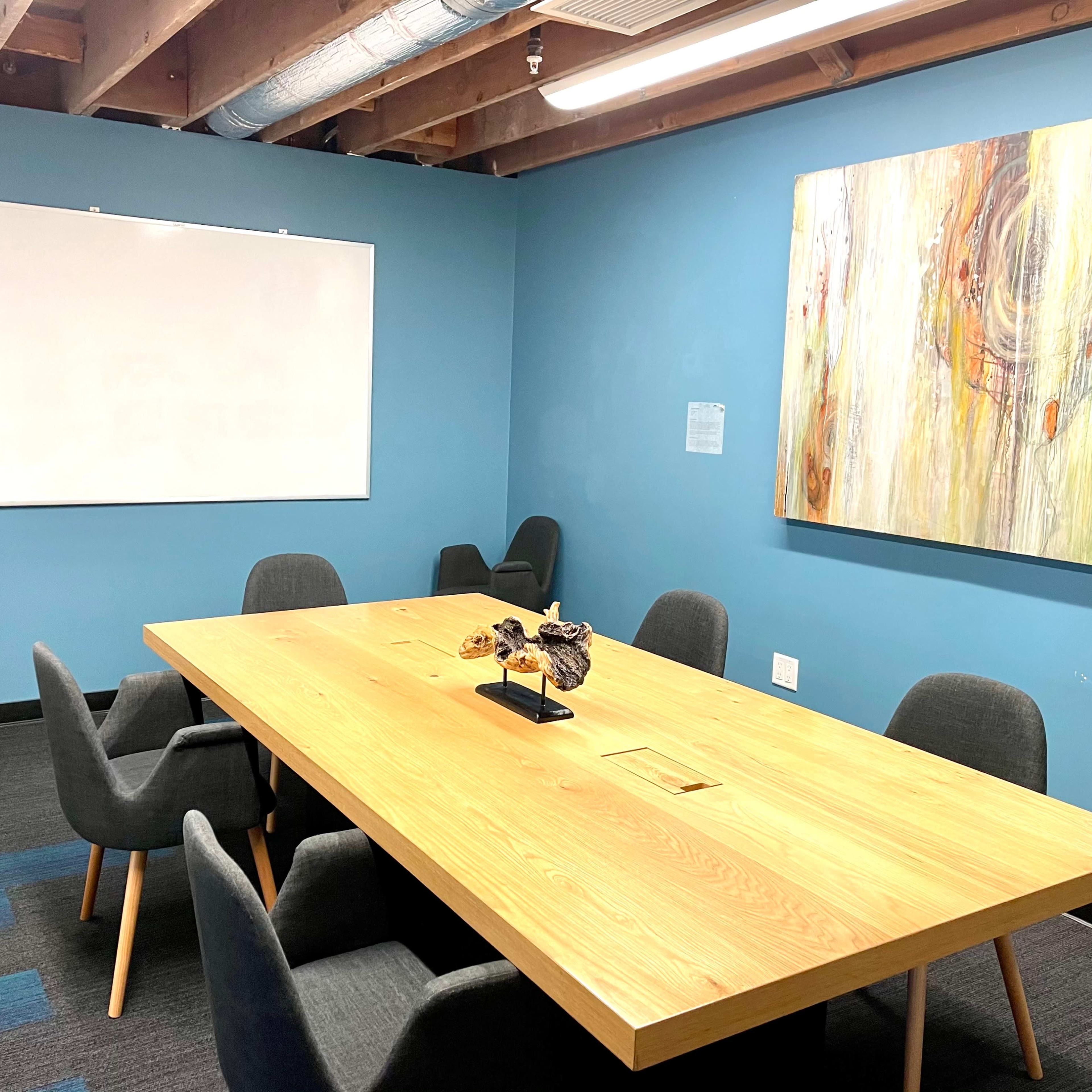 A modern conference room with a large wooden table surrounded by gray chairs, a whiteboard on the wall, and a colorful abstract painting.