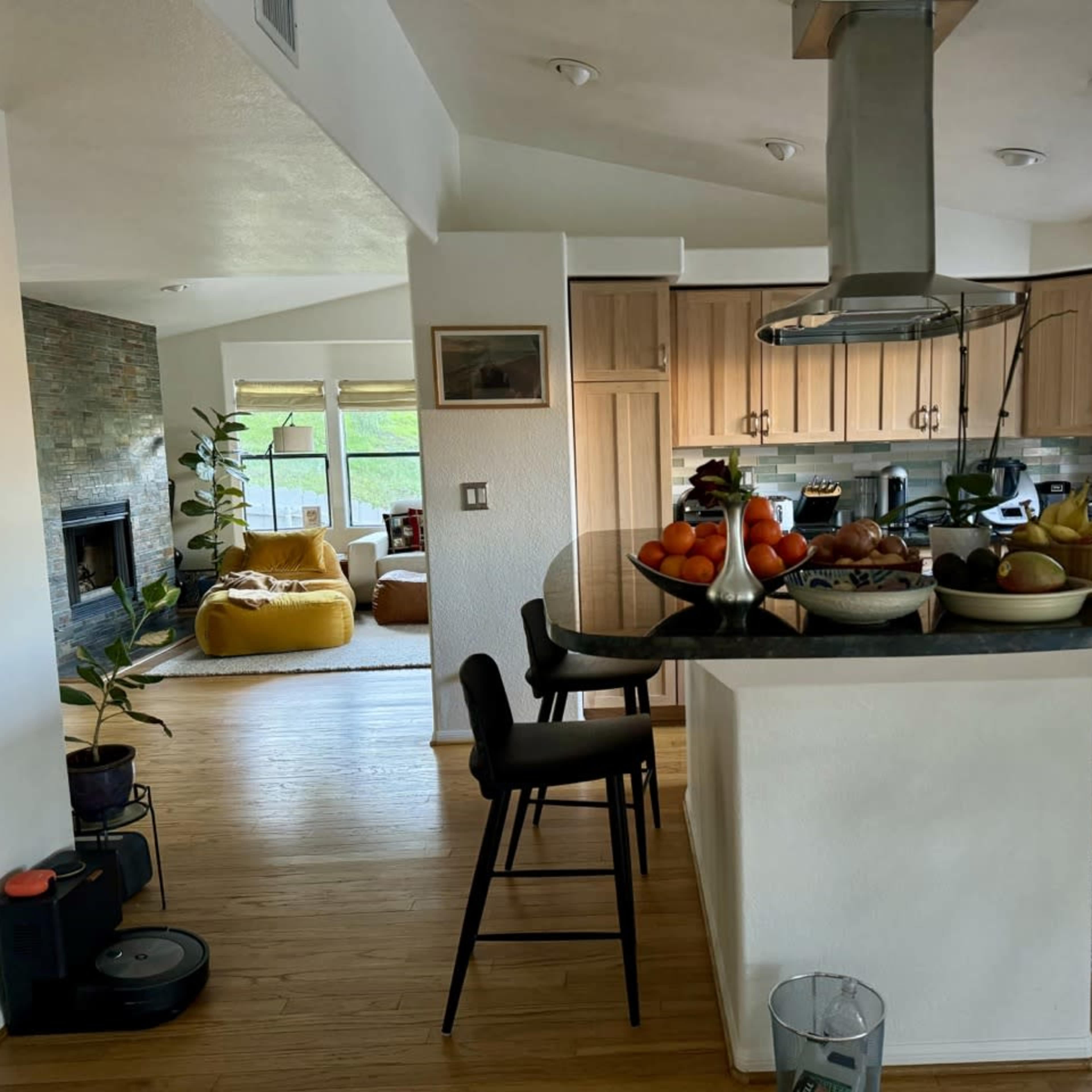 A modern kitchen features a central island with a large bowl of fruit, overlooking a living area with a cozy seating arrangement and large windows.