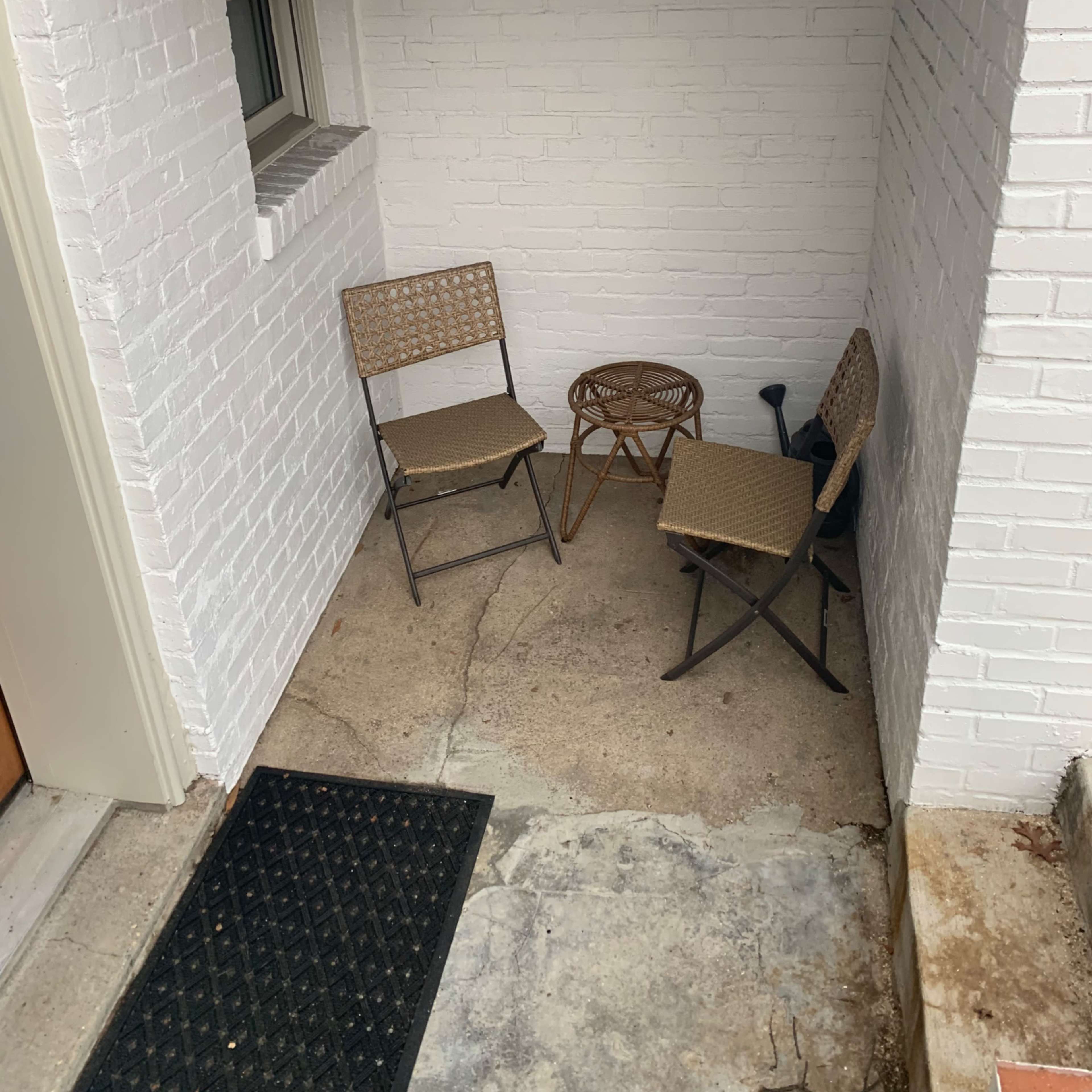 A small, corner seating area with two metal chairs and a round wicker table, situated against a white brick wall.