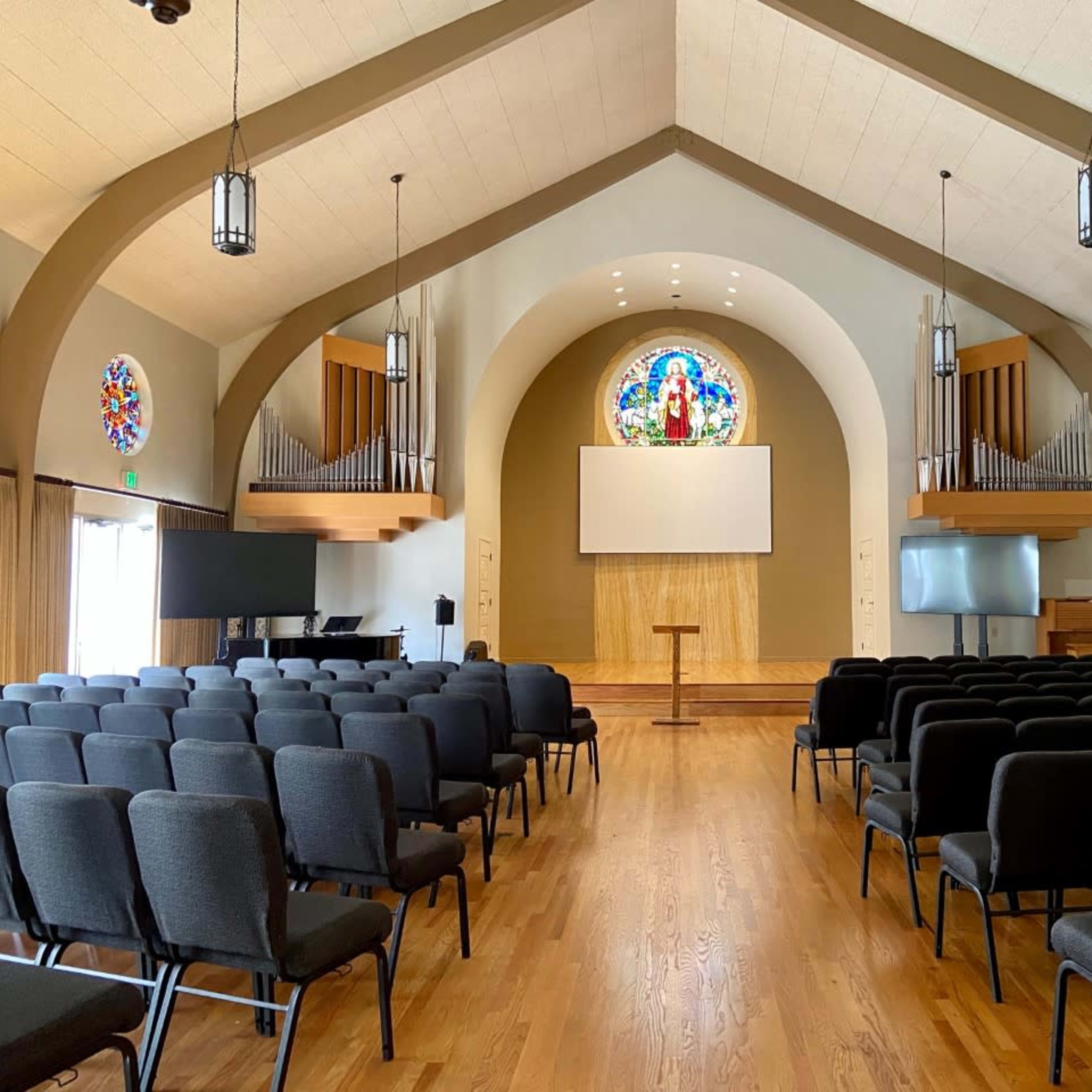 A spacious church interior with rows of black chairs facing a wooden podium and a large screen, flanked by stained glass windows and organ pipes.