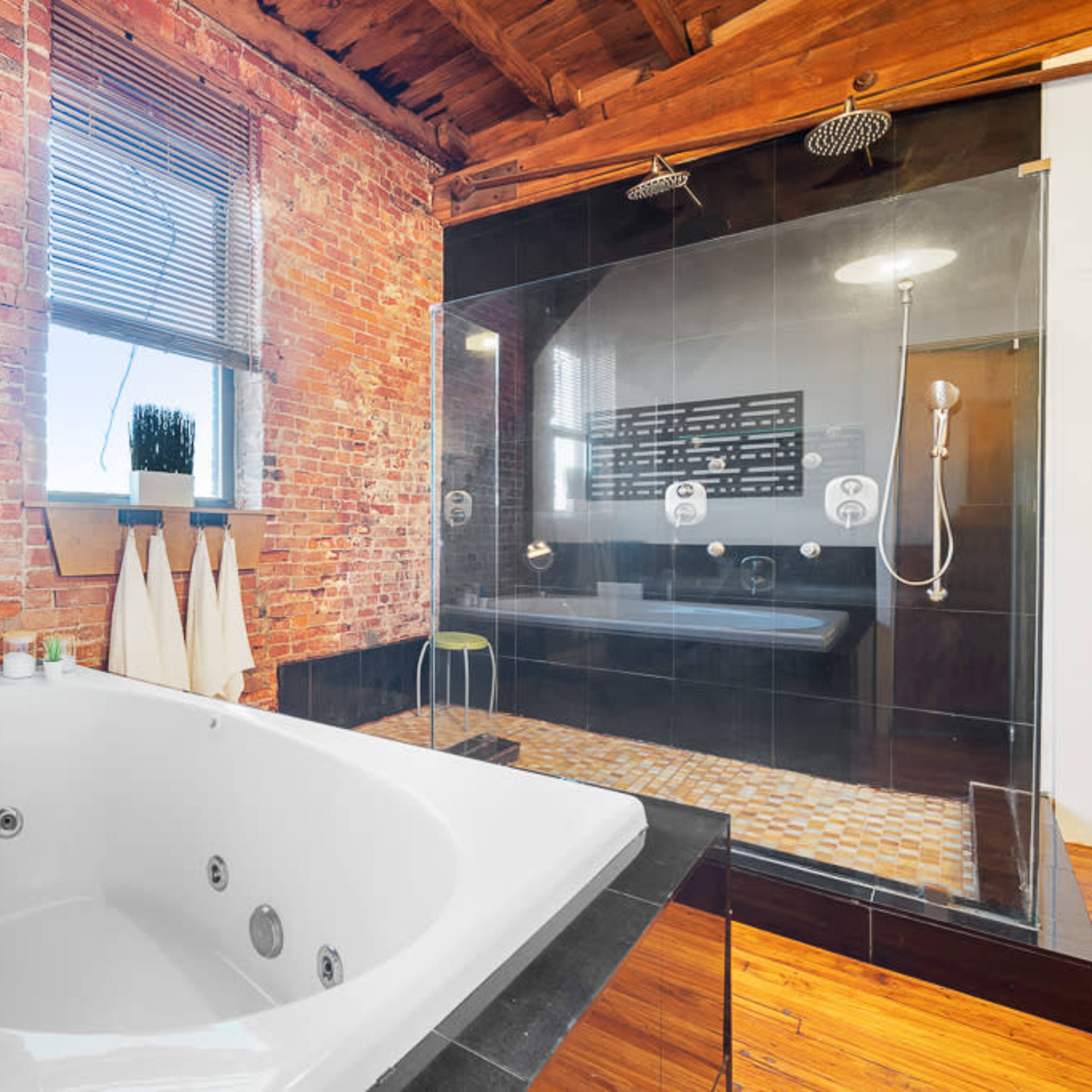 A modern bathroom with a large white jacuzzi tub and a glass-enclosed shower area, complemented by exposed brick walls and wooden beams.