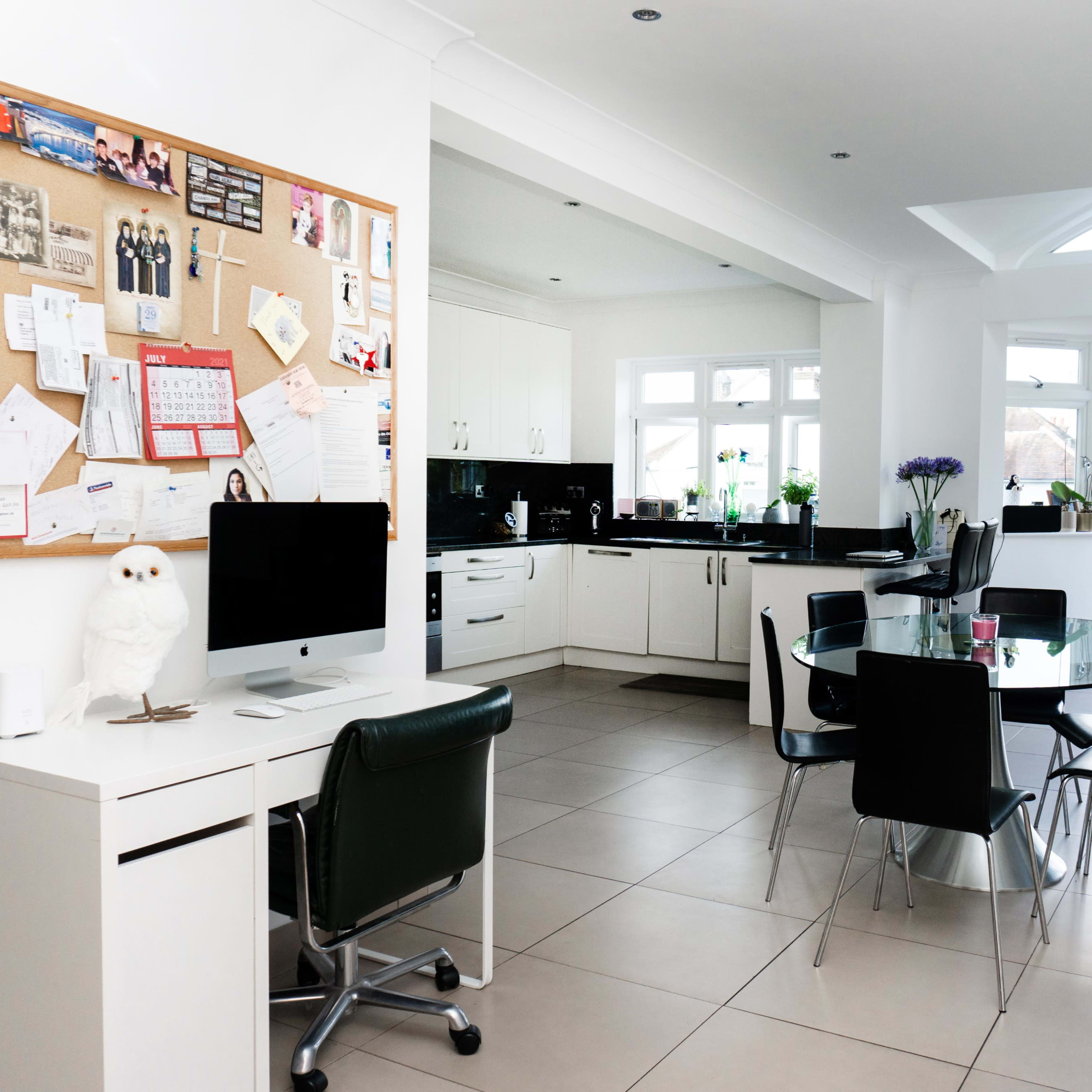 A modern kitchen and dining area features a bulletin board, a computer setup, a circular dining table, and large windows that provide natural light.