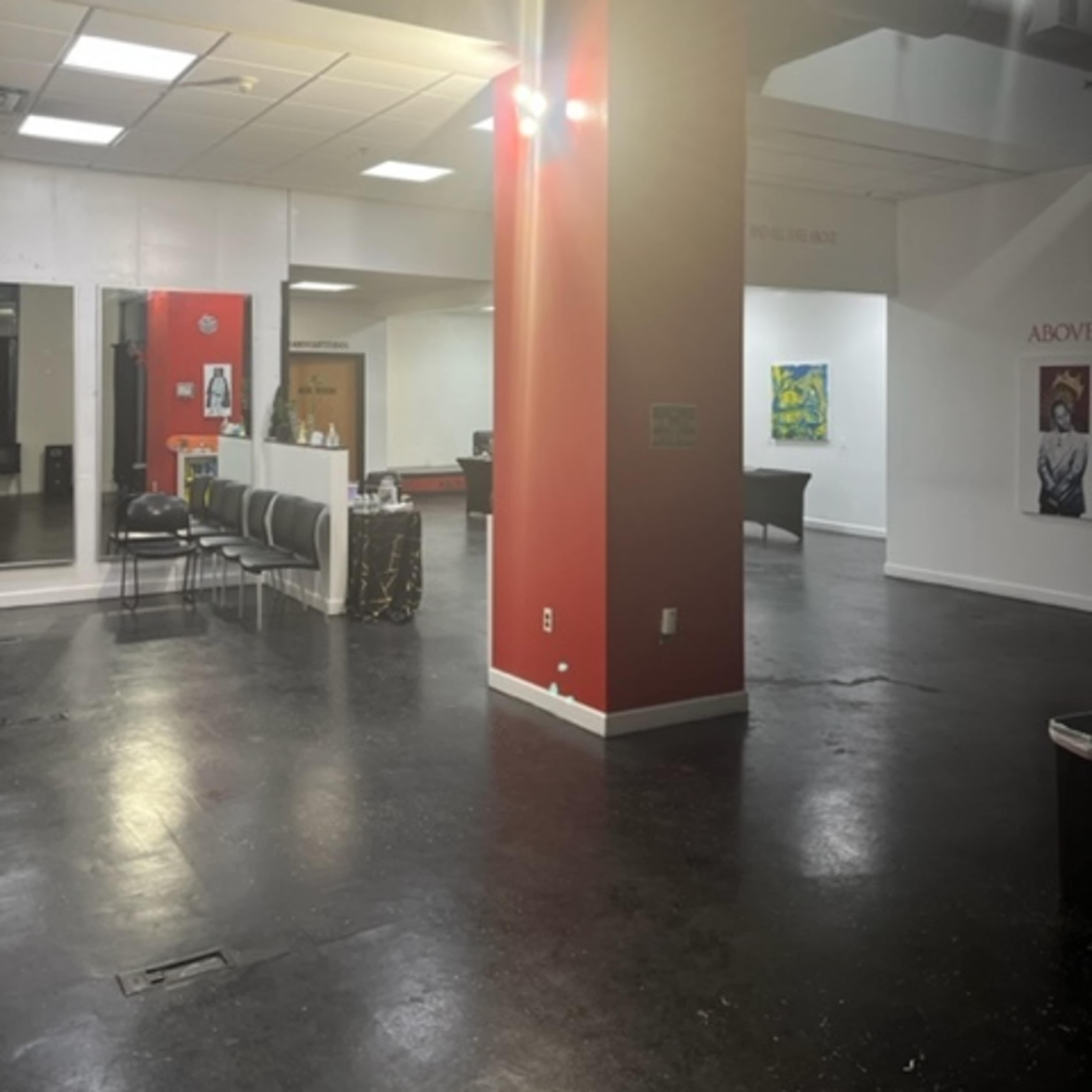 A spacious, minimalistic interior with red columns, black flooring, and a few scattered furniture pieces, including chairs and a piano.