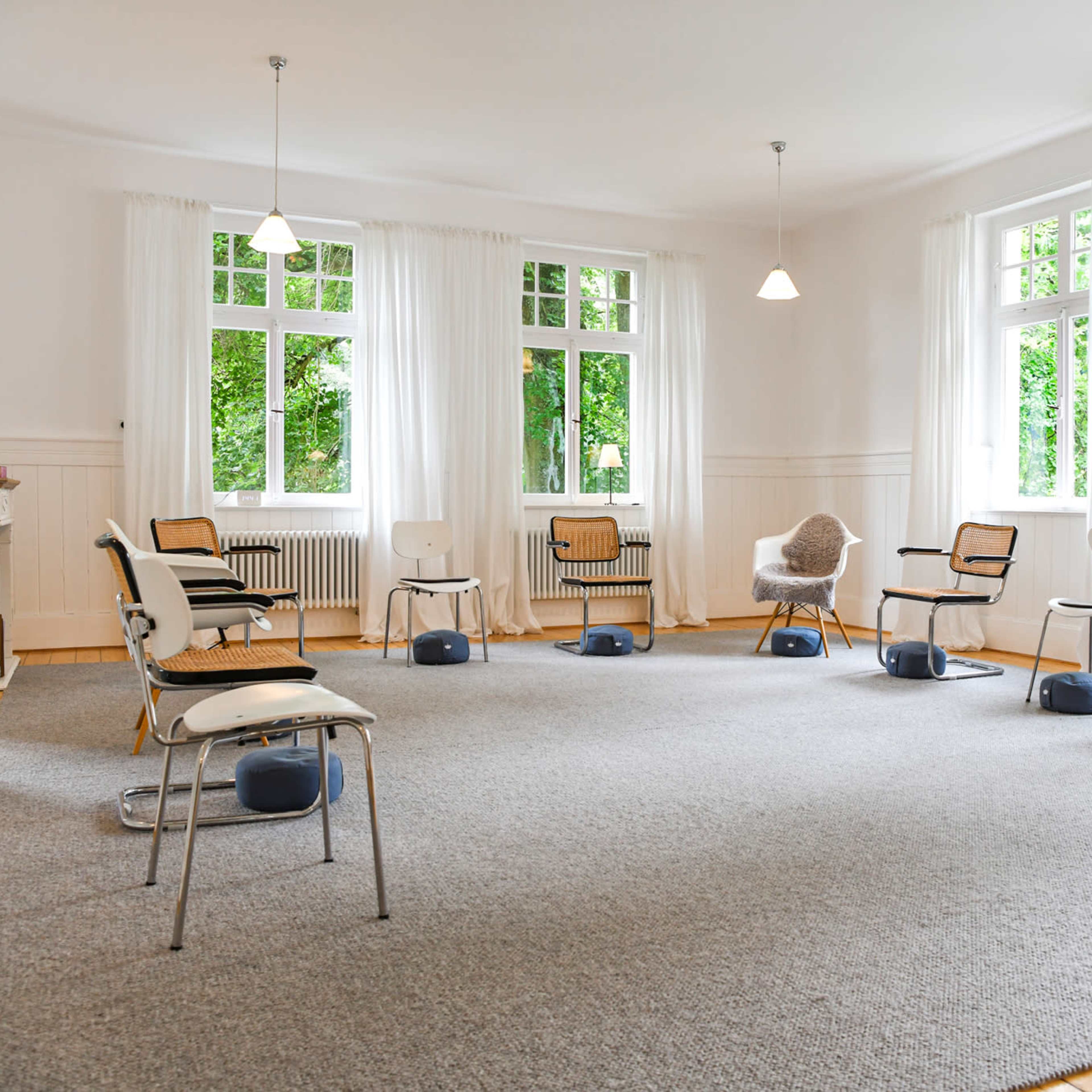 A bright, spacious room features several chairs arranged in a circle on a carpeted floor with large windows allowing natural light in.