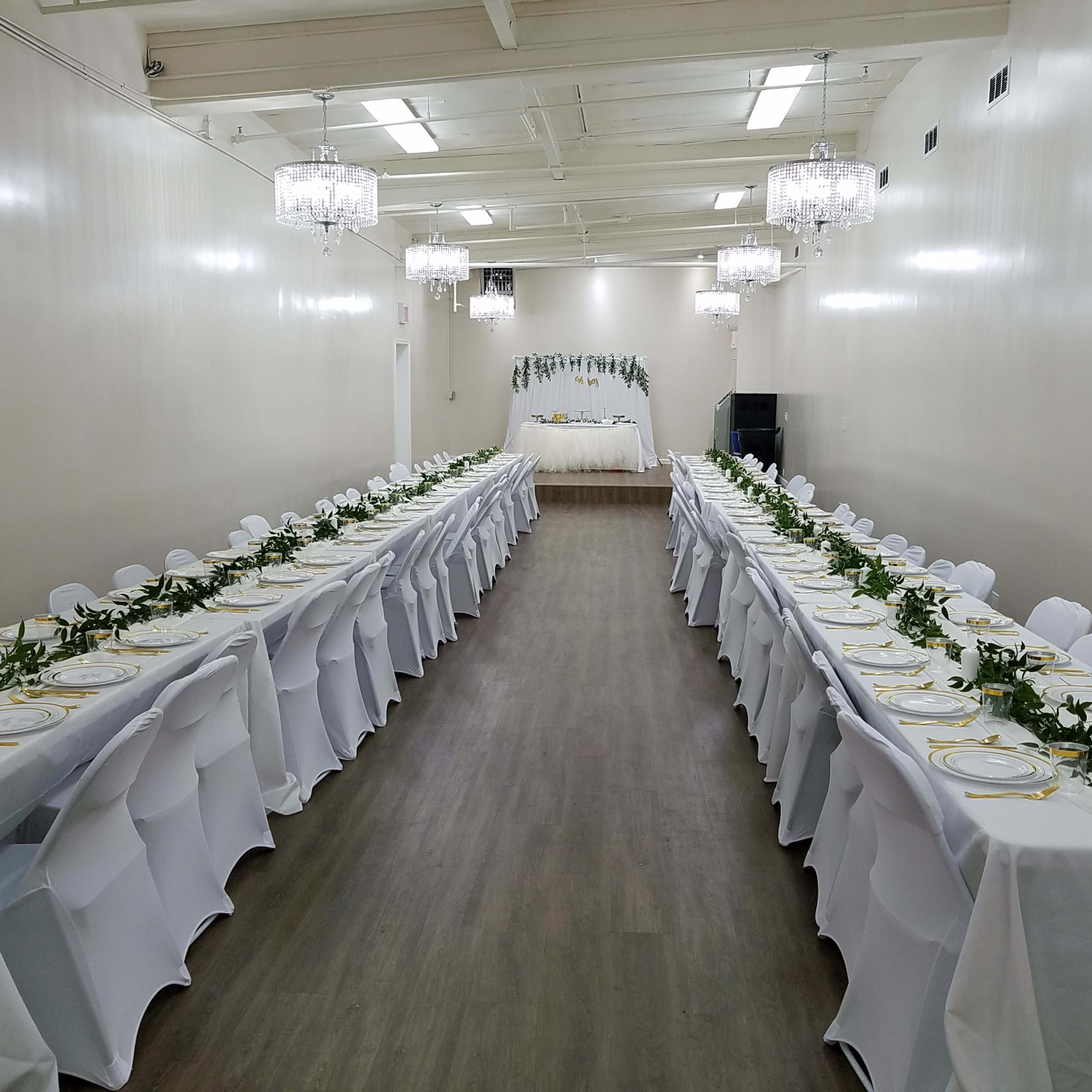 A long banquet hall with two rows of elegantly set tables featuring white tablecloths and greenery decorations, leading to a decorated stage at the far end.