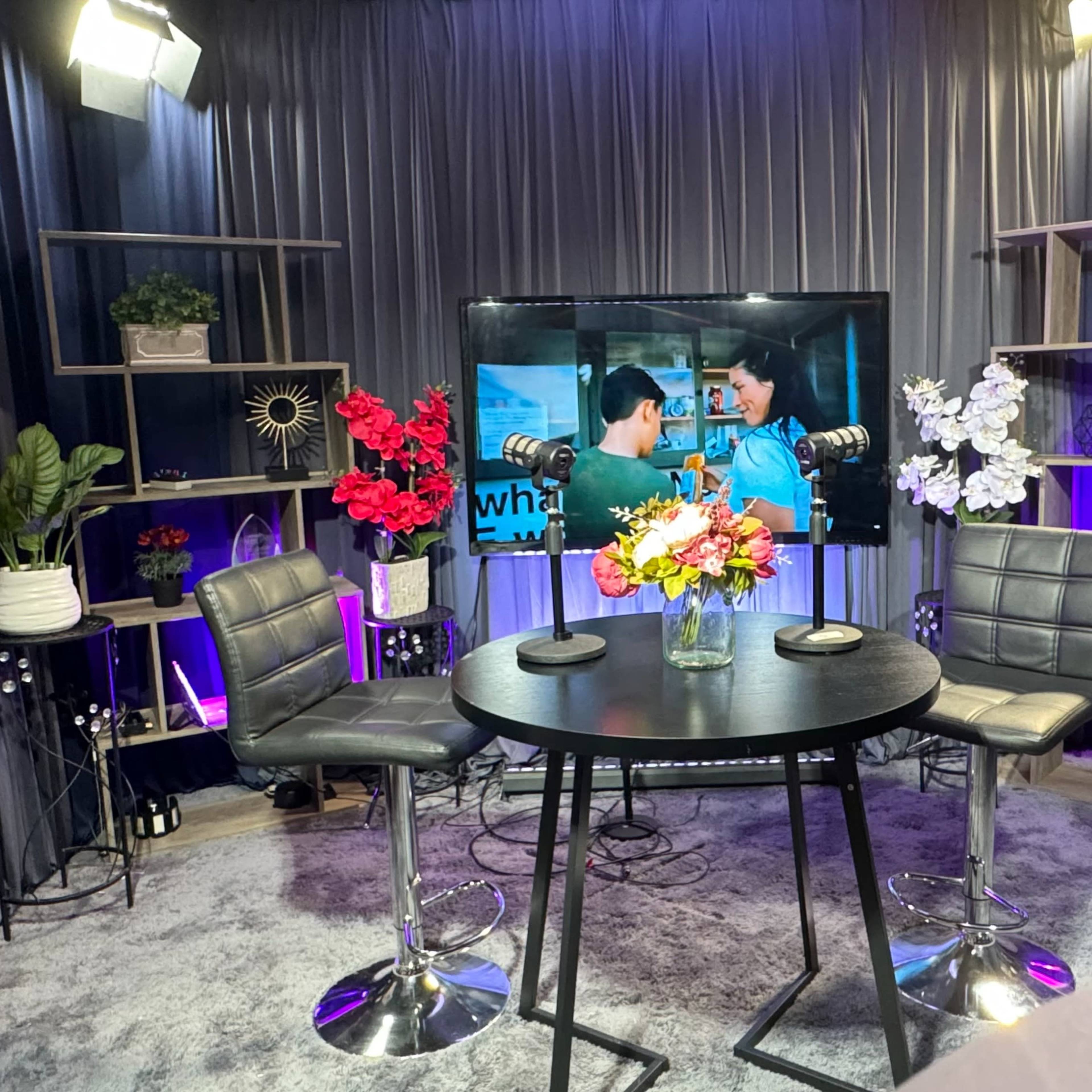A television studio setup with a round table, two black chairs, a large screen displaying a video, and various potted plants and flowers arranged on shelves.