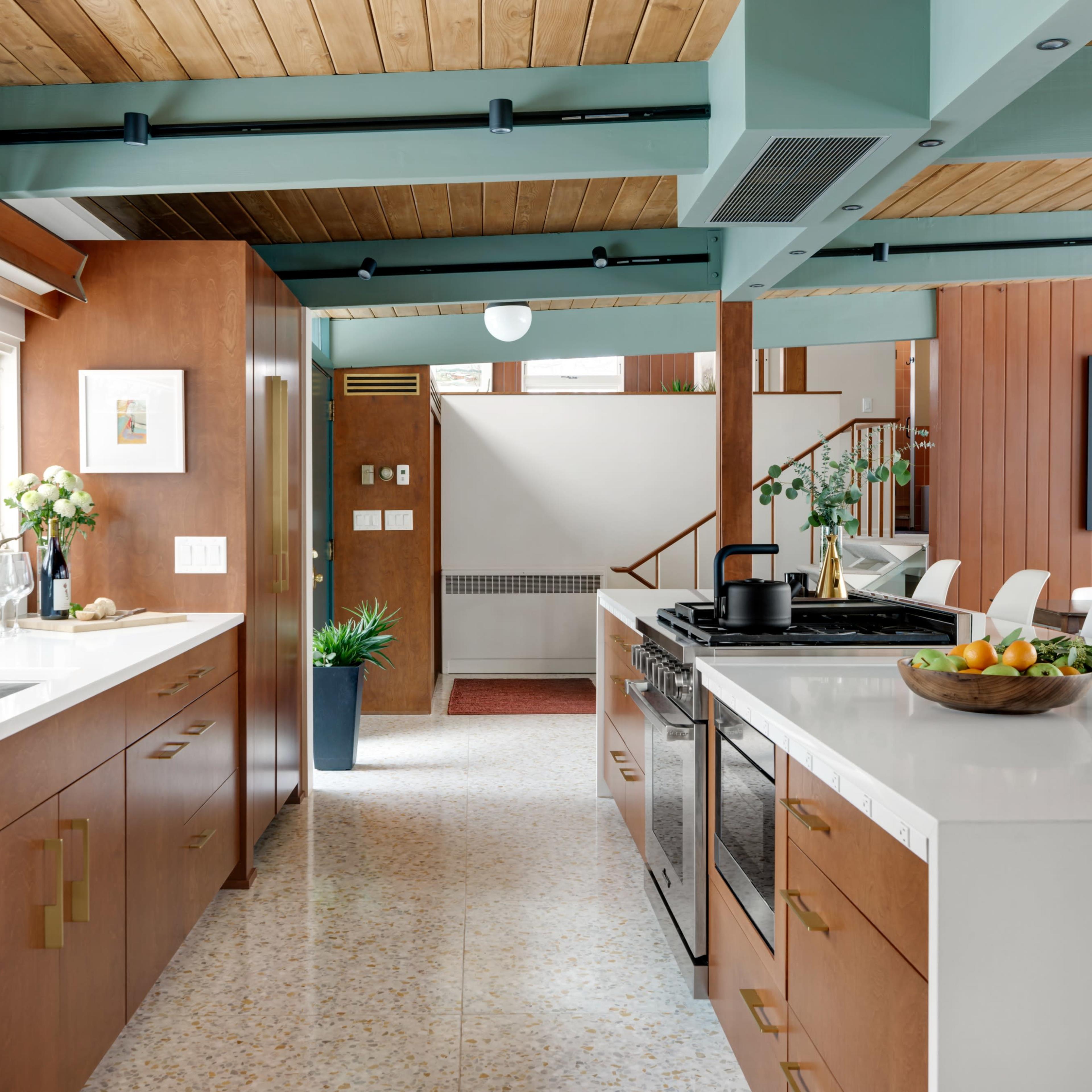 A modern kitchen with wooden cabinetry, white countertops, and a view into a dining area featuring a staircase and colorful decor.