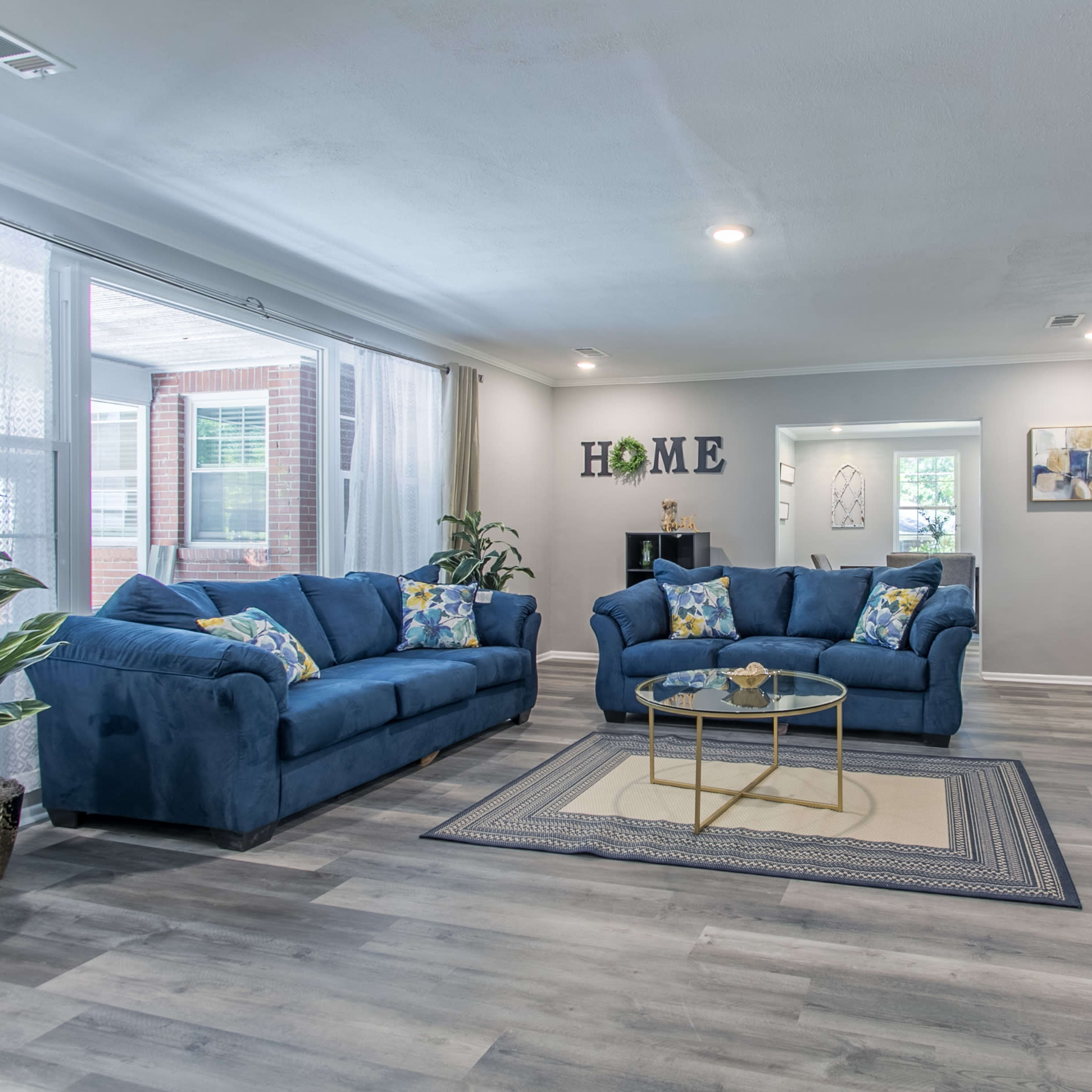 The living room features two blue sofas with decorative pillows, a glass coffee table, and a fireplace, with light streaming through large windows.