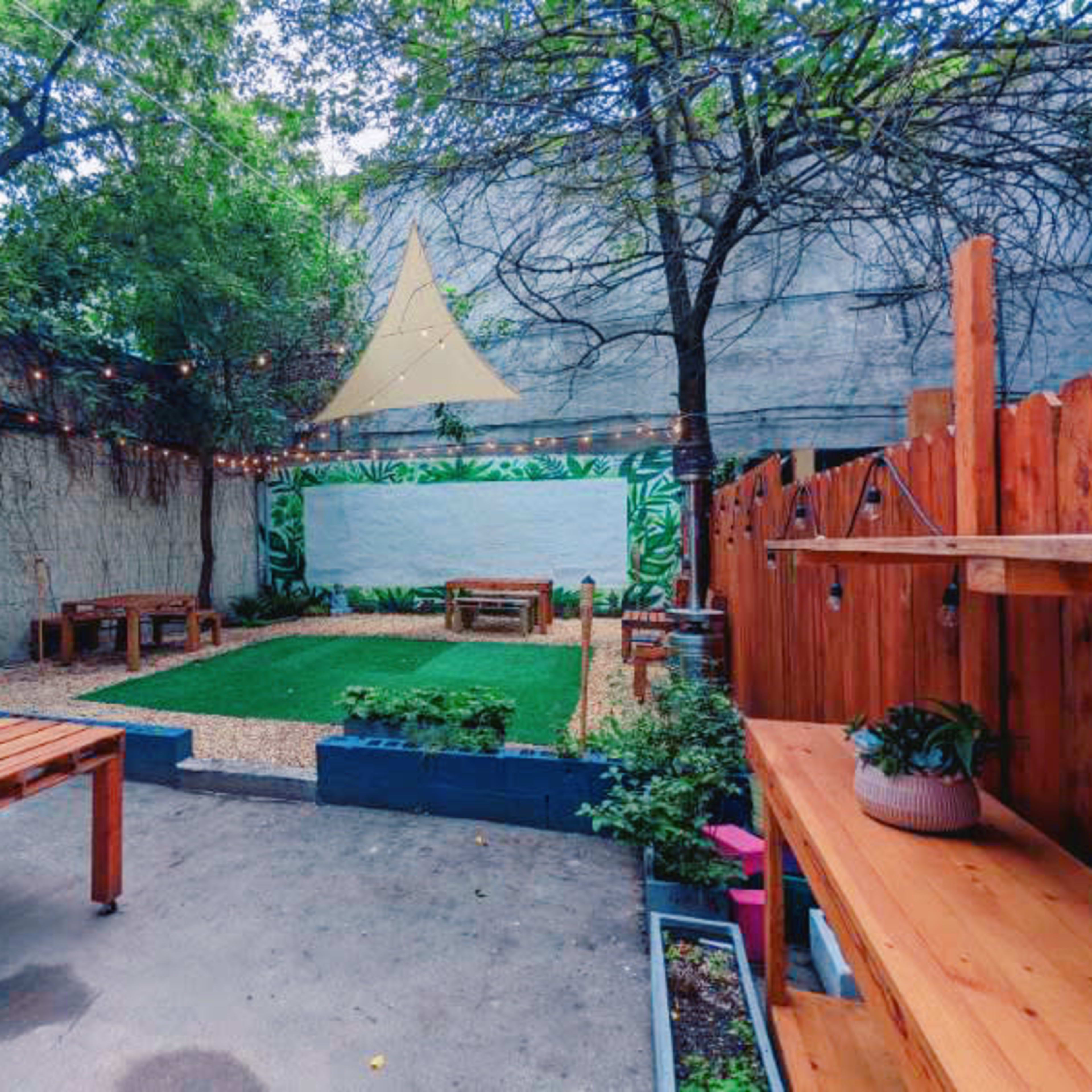 A backyard space featuring wooden benches, a grassy area, and a large white wall surrounded by greenery and string lights.