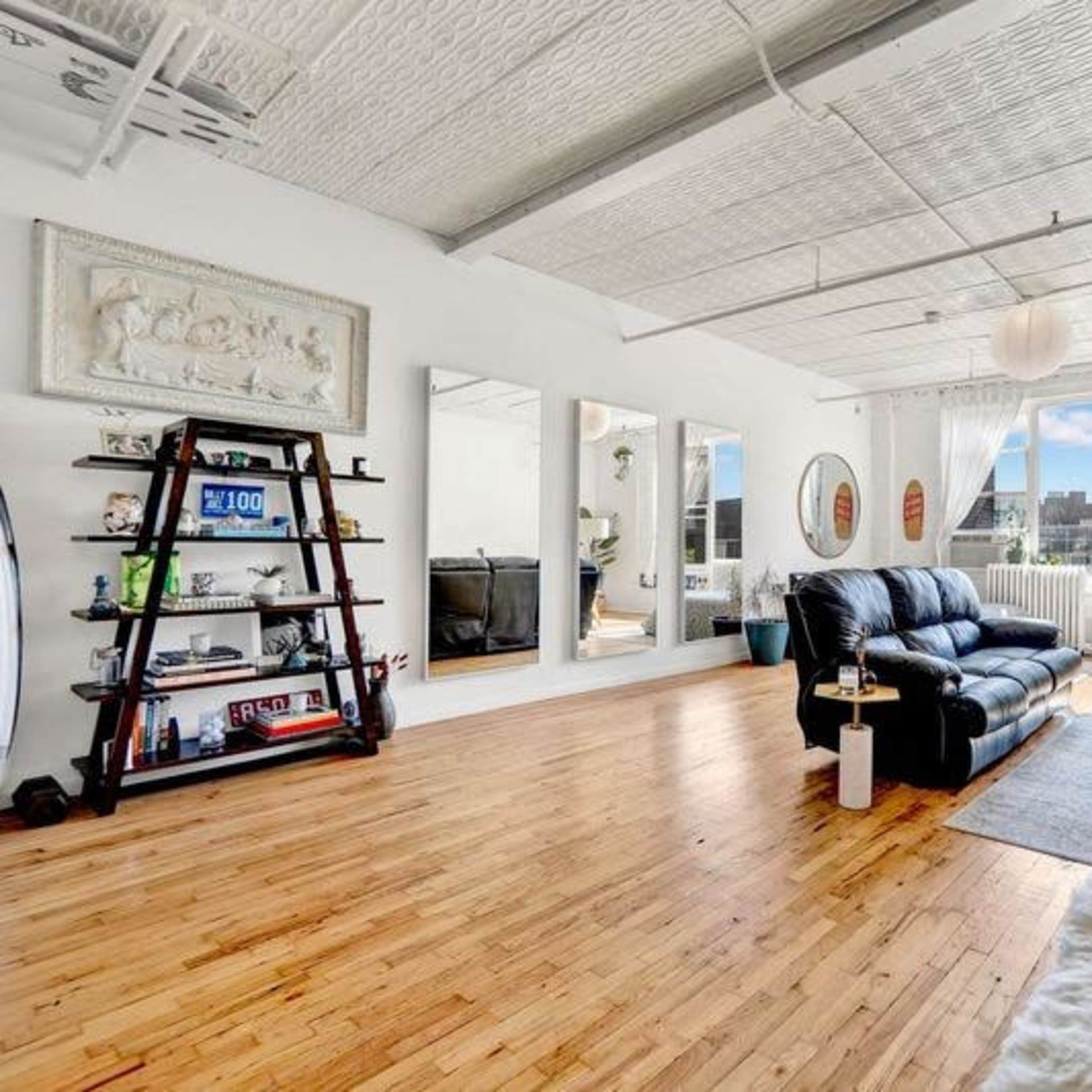 A spacious living room with a black leather sofa, wooden flooring, and large windows allowing natural light to enter, complemented by decorative mirrors and plants.