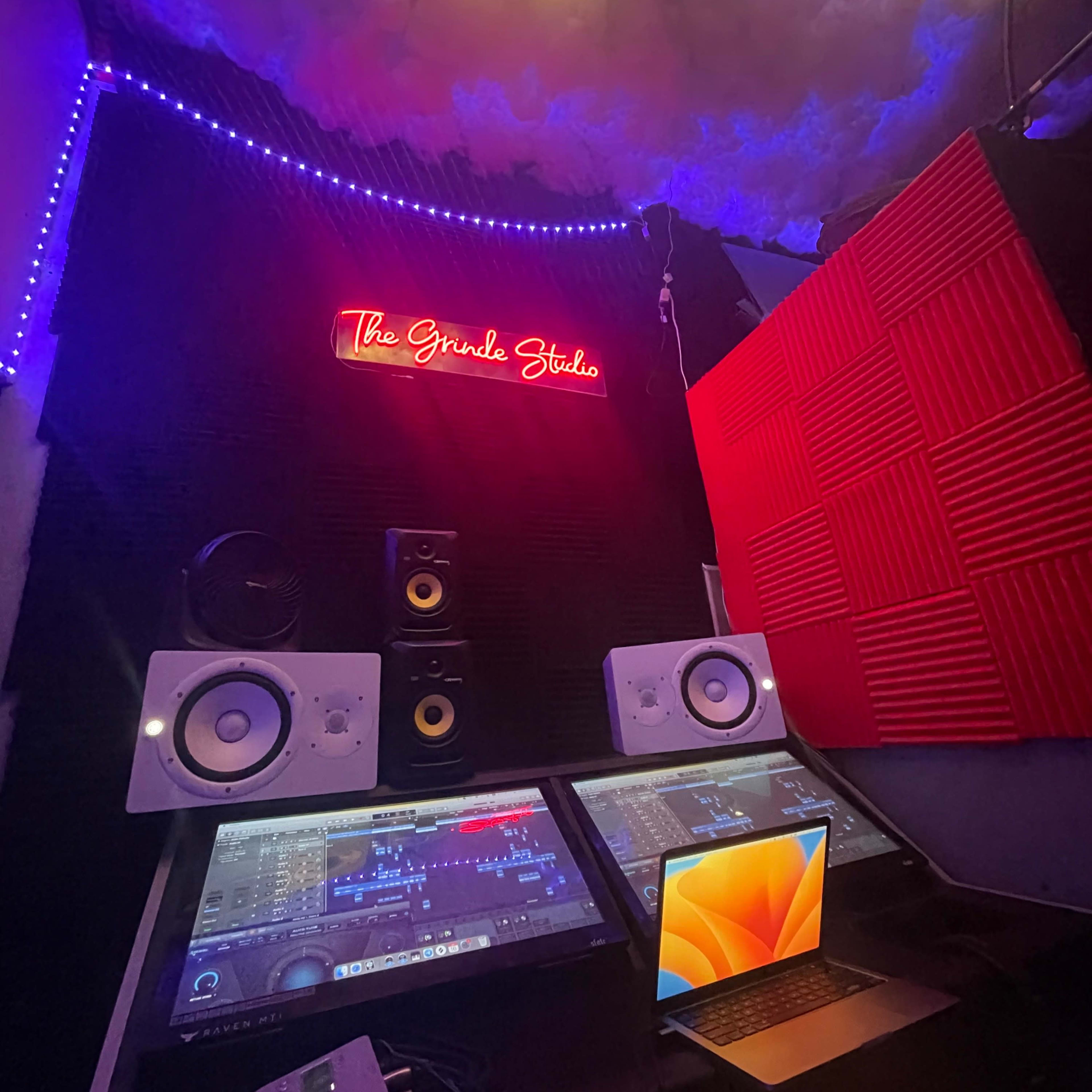 A music production studio featuring two monitors, speakers, and an illuminated sign that reads "The Grind Studio."