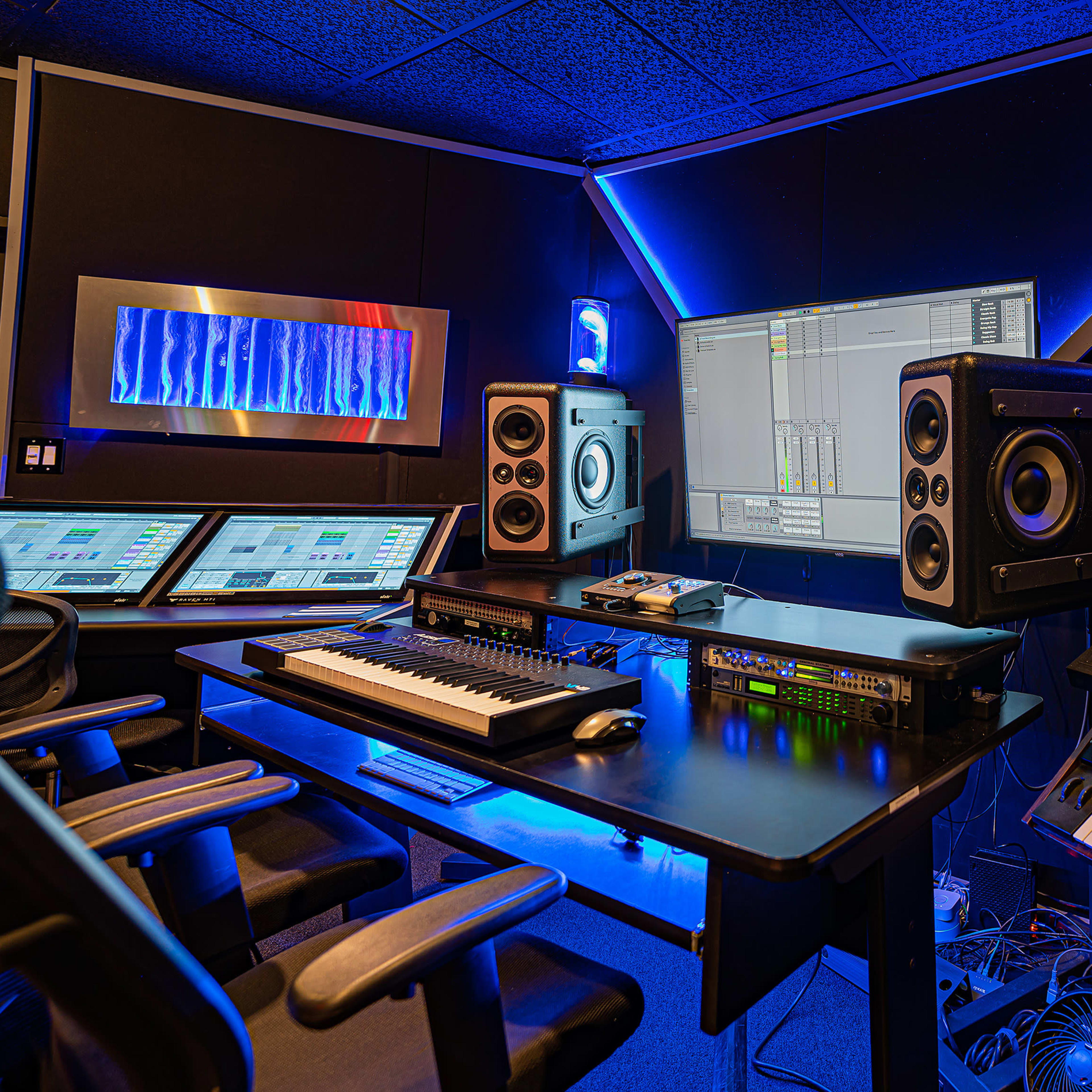 A modern music production studio equipped with a computer, mixing consoles, speakers, and multiple keyboards surrounded by blue lighting.