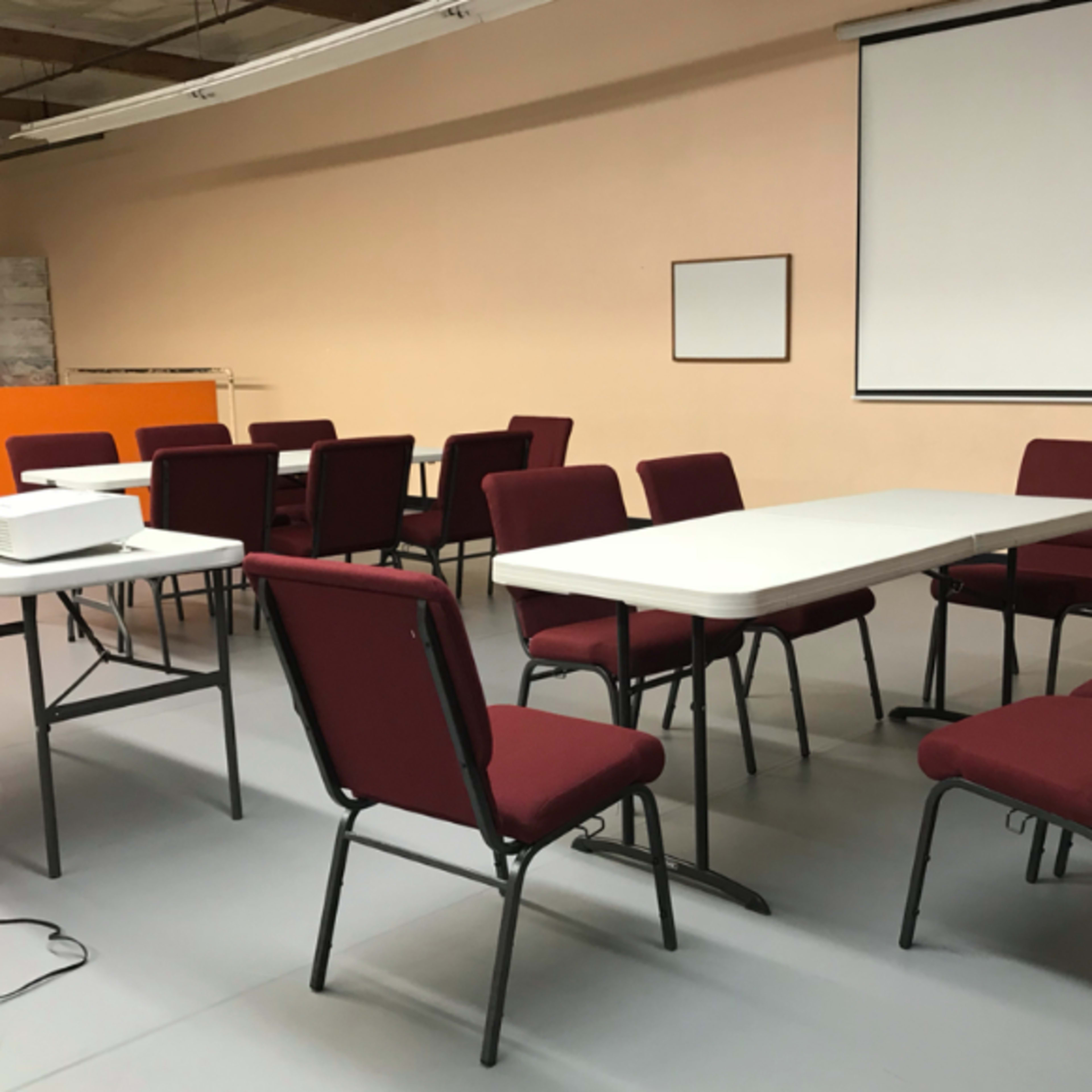 A meeting room is arranged with several chairs and tables, featuring a projector and a blank screen.