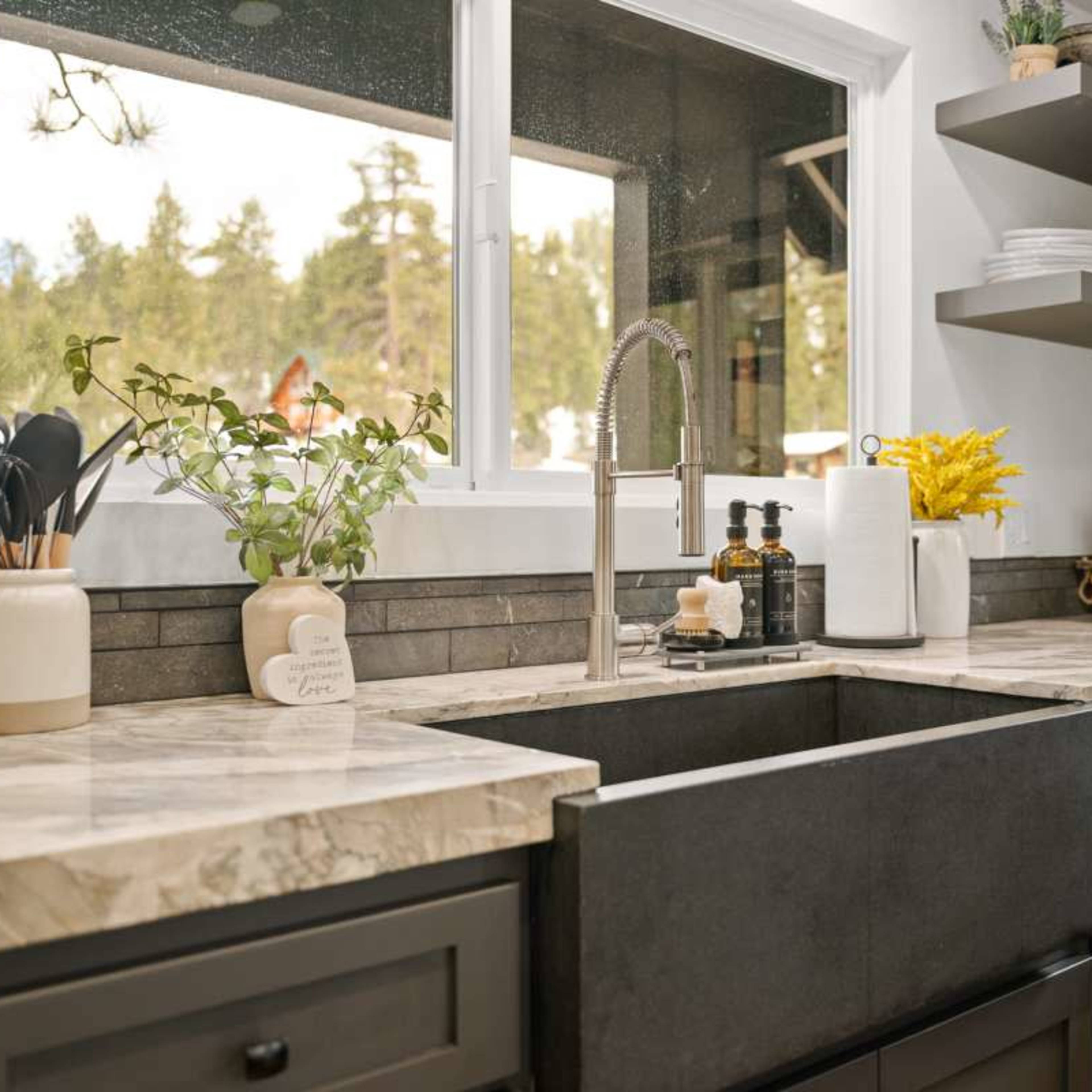 A modern kitchen with a large farmhouse sink, marble countertops, and open shelves displaying dishes and greenery.