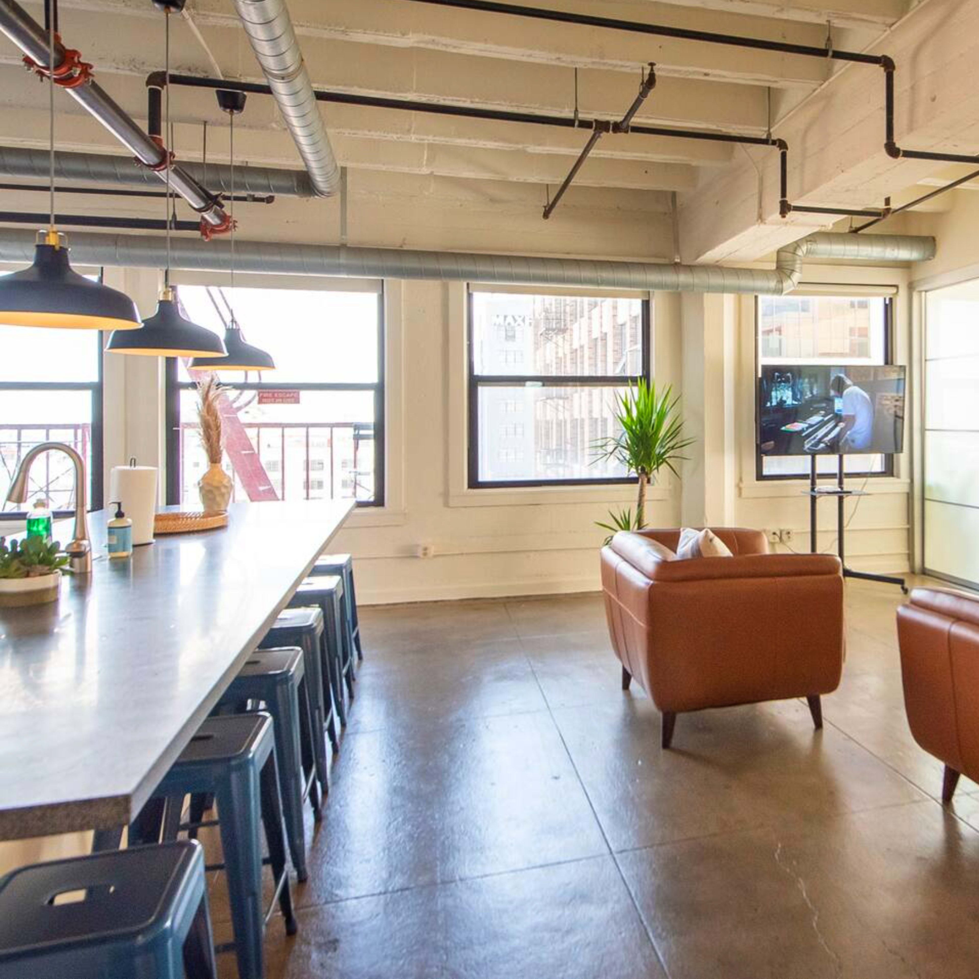 A modern office lounge area with two brown leather chairs, a long table with metal stools, and a television facing the seating.