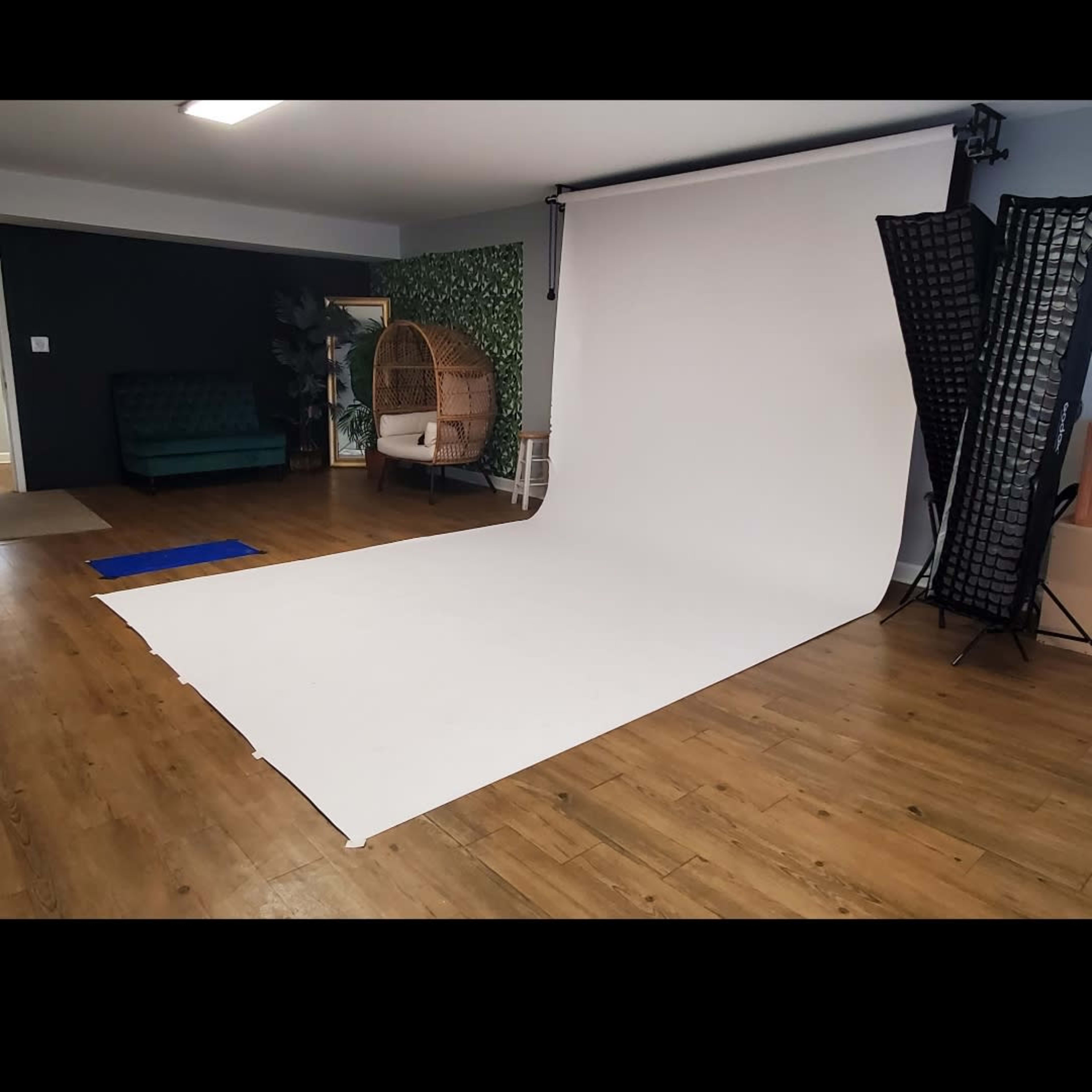 A photography studio with a white backdrop, wooden floor, a green couch, a decorative plant wall, and lighting equipment on the side.