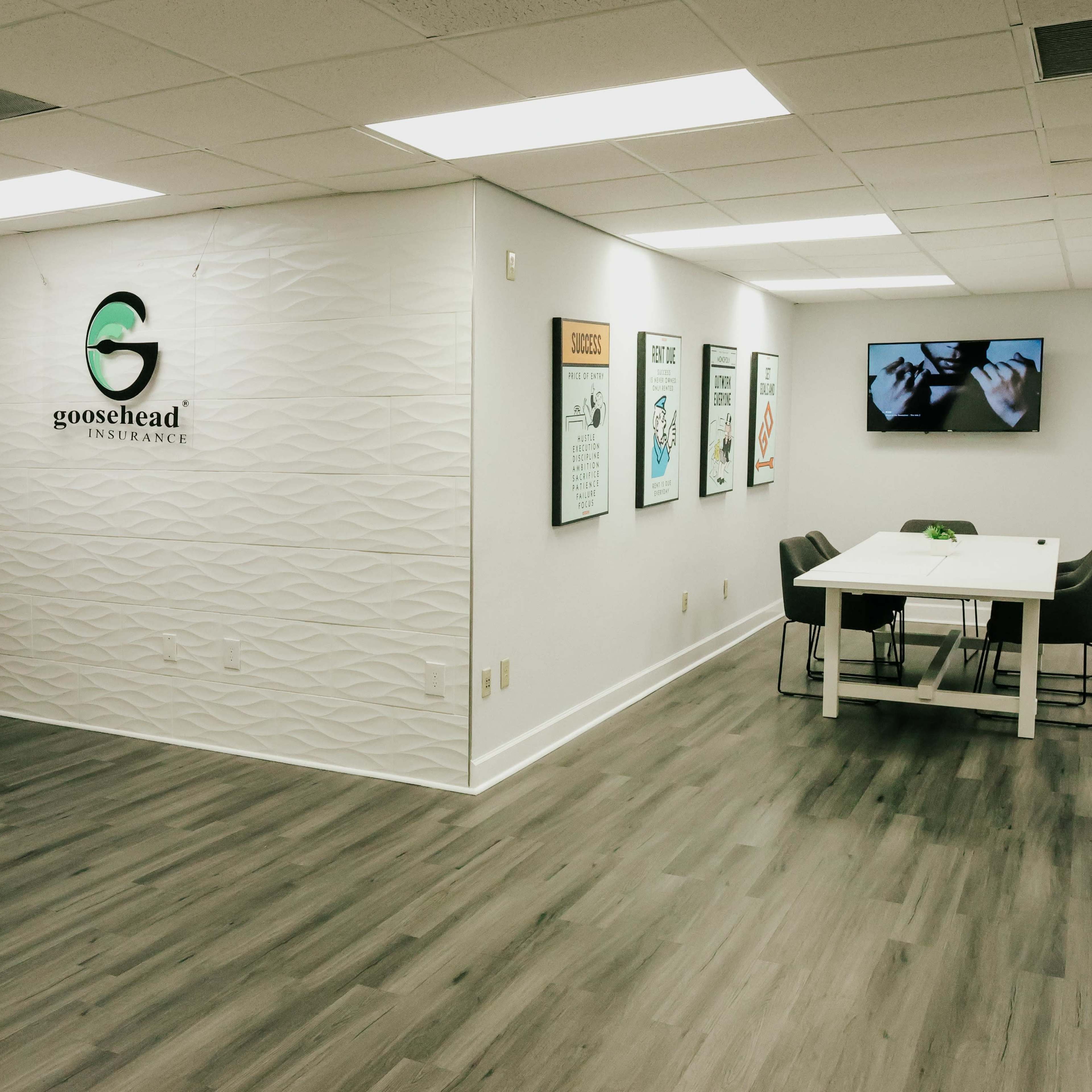 A modern office of Goosehead Insurance, featuring a desk area on the left, a textured wall with the logo, and a meeting space with a table and chairs on the right.