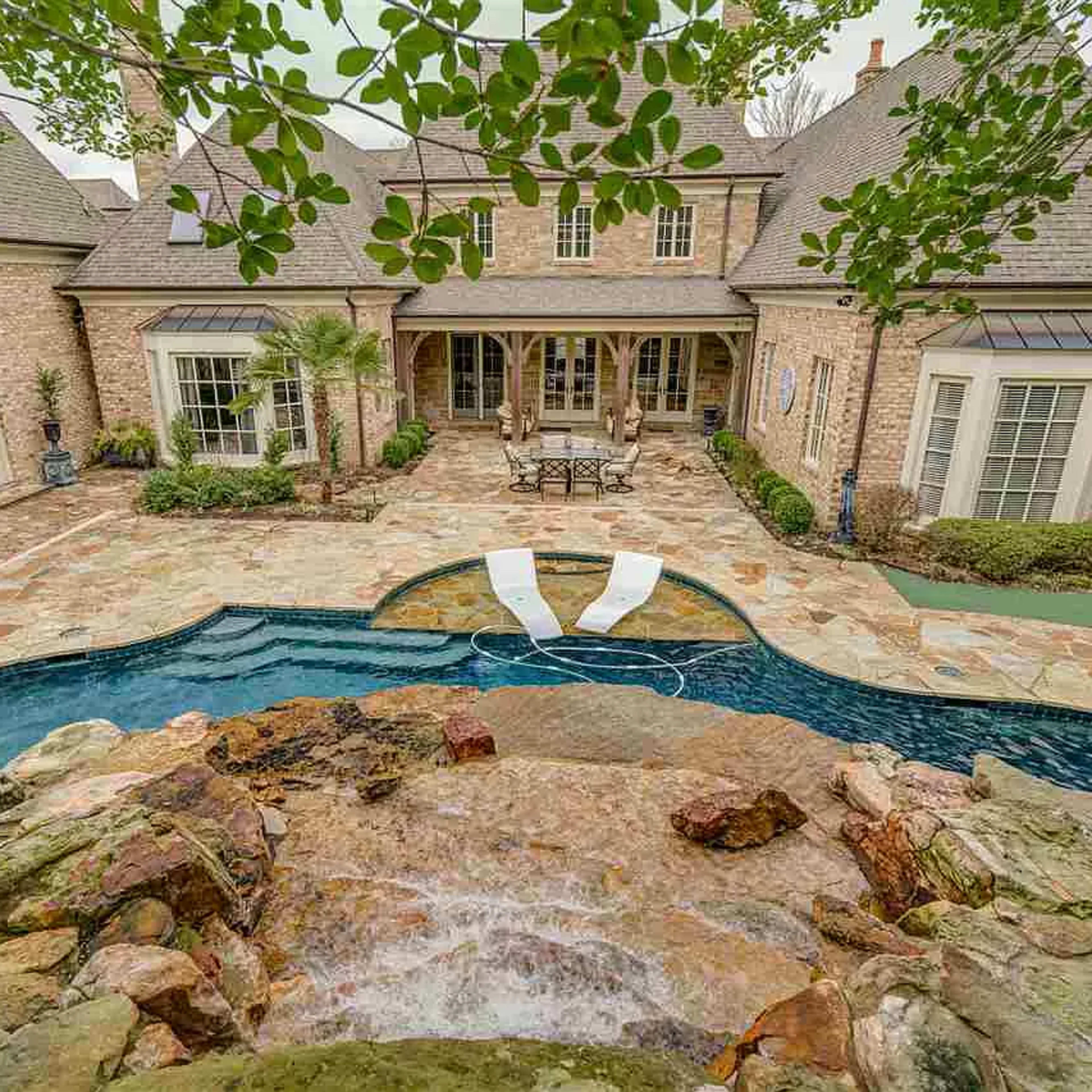 A stone patio surrounds a swimming pool with a waterfall feature and lounge chairs, framed by the exterior of a large brick house and landscaped greenery.