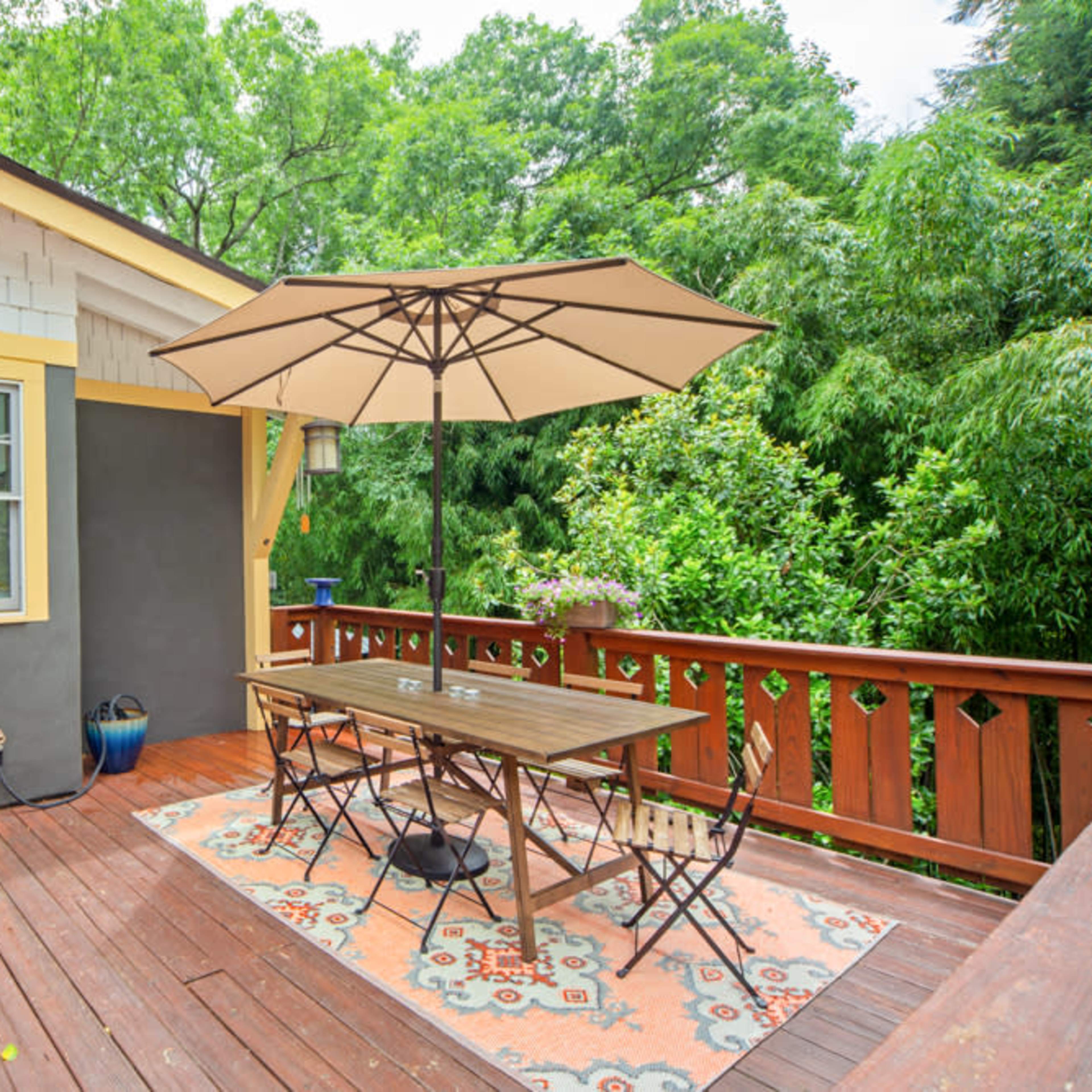 A wooden deck features a table and chairs under a large patio umbrella, surrounded by green trees and potted plants.