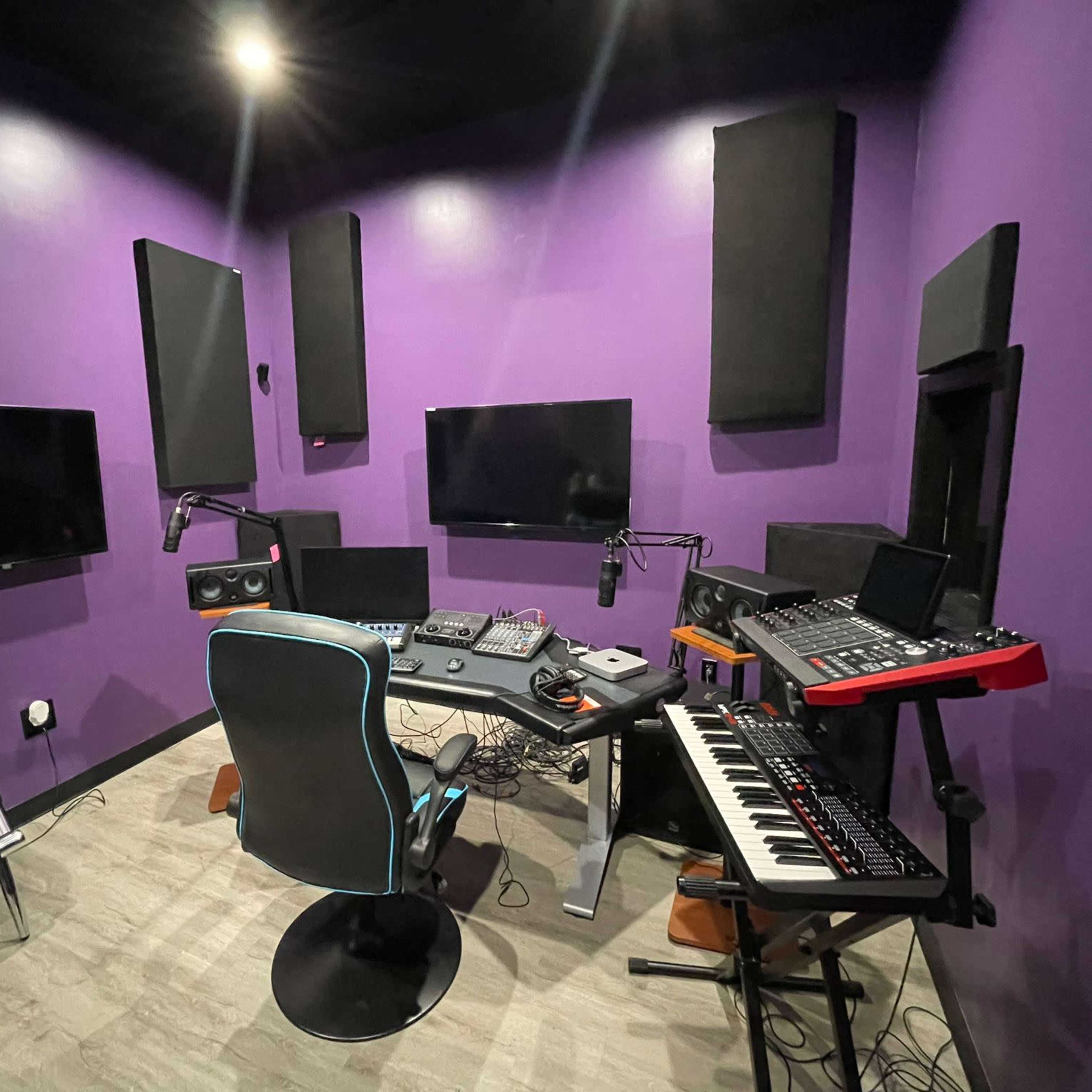 A small music production studio features a desk with various audio equipment, a keyboard, and multiple monitors against a purple wall.
