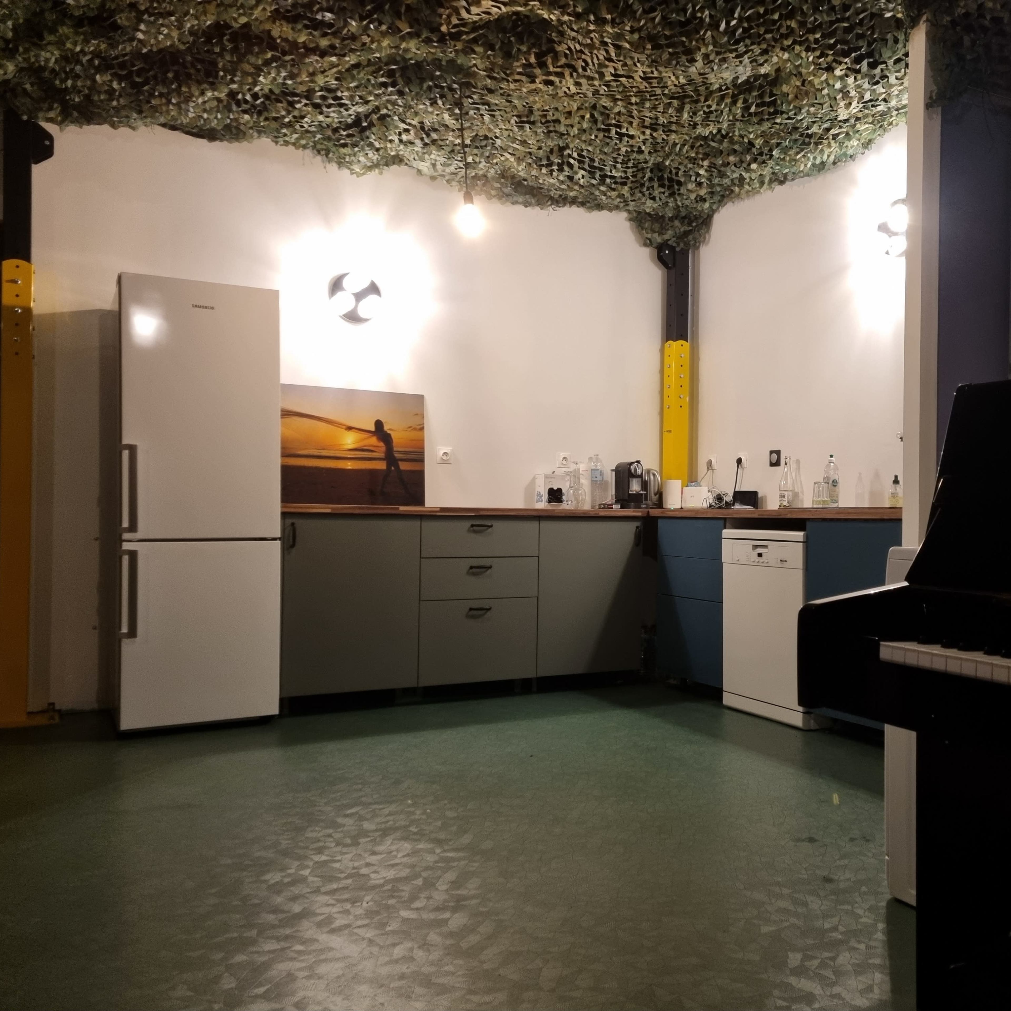 A modern kitchen with a white refrigerator, gray cabinets, a blue drawer unit, and a black piano in a room with a camouflage ceiling.