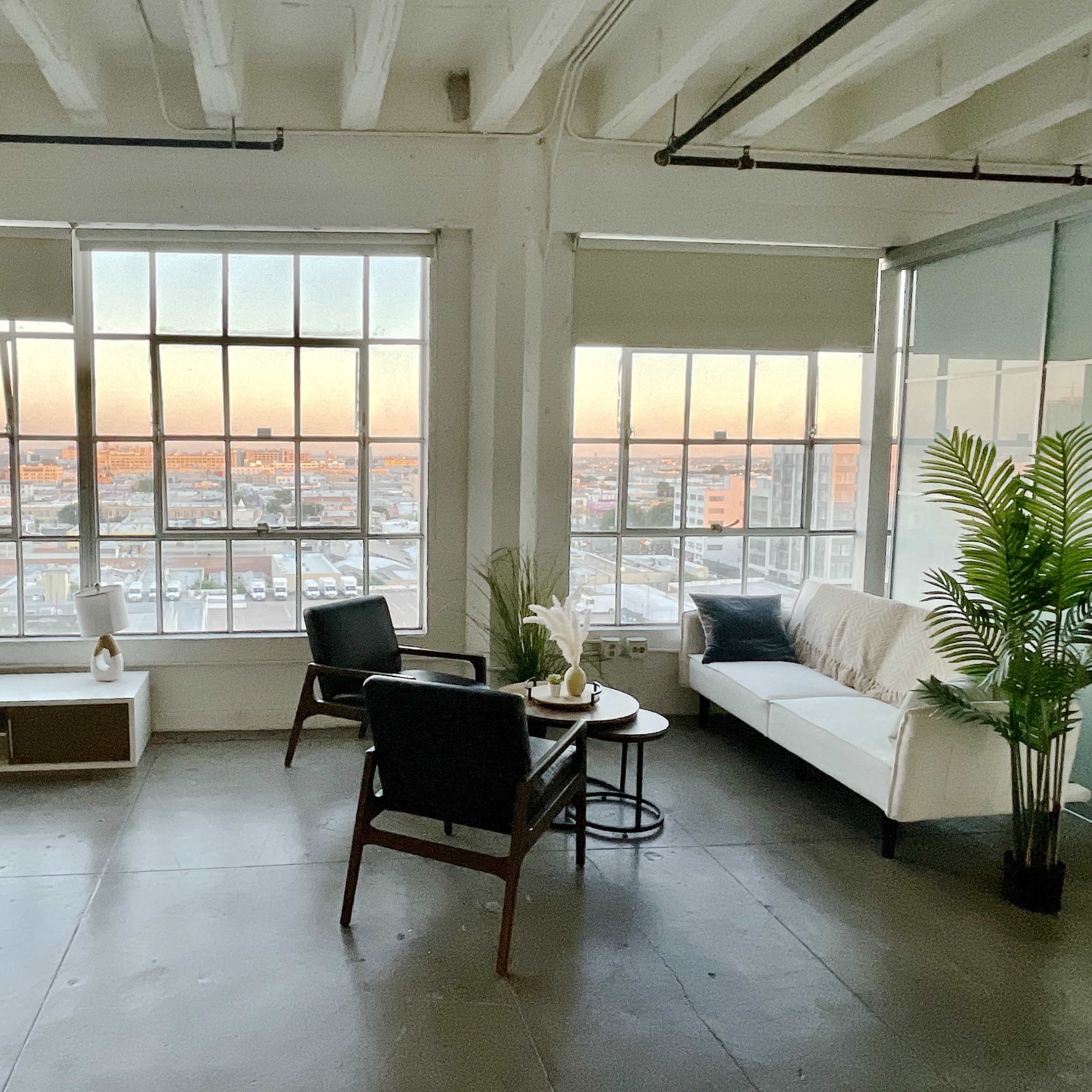 A modern, airy interior with large windows providing a view of a cityscape at sunset, featuring minimalist furniture including a sofa, chairs, and a coffee table.