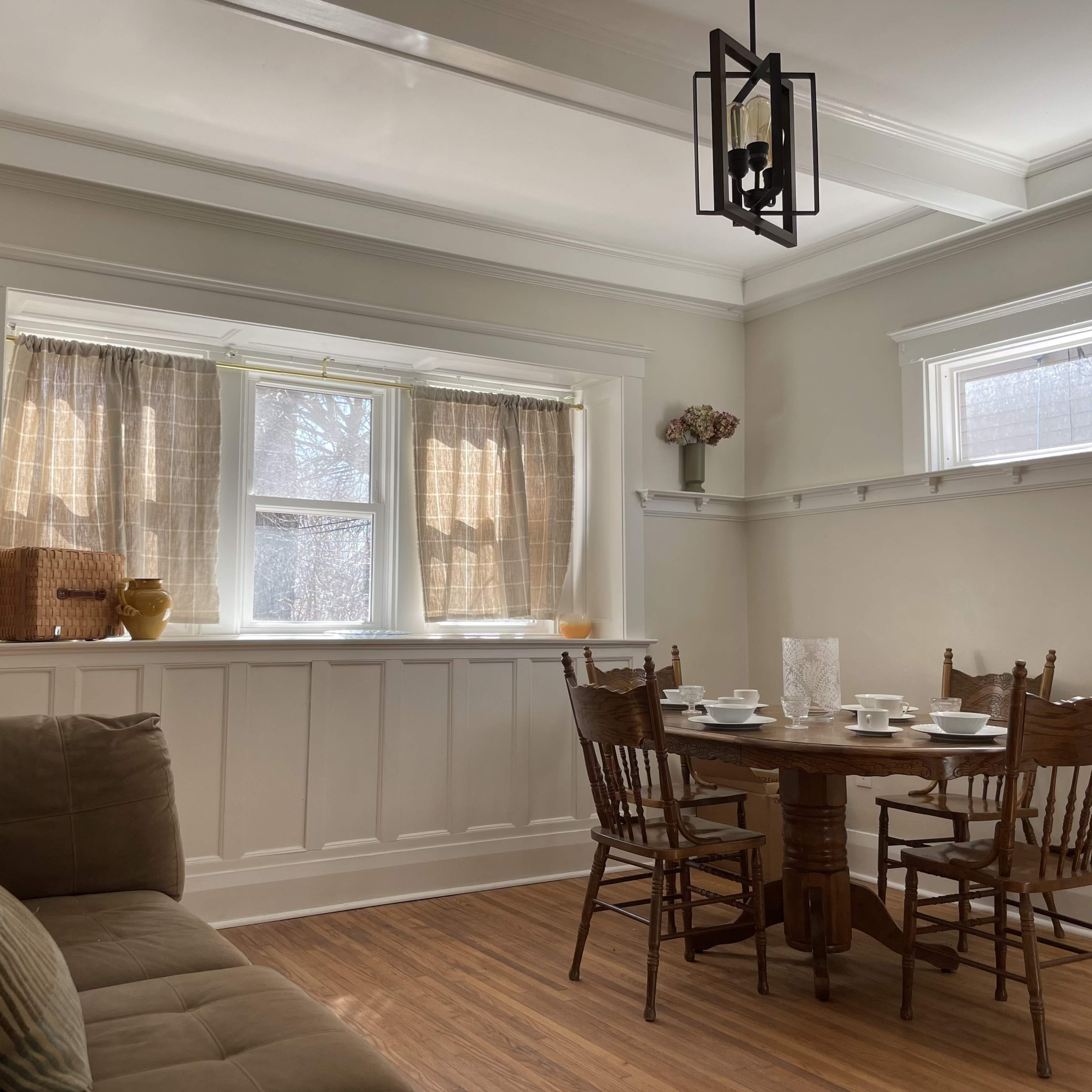 A dining area features a round wooden table set for four, surrounded by chairs, with a couch and window curtains in the background.