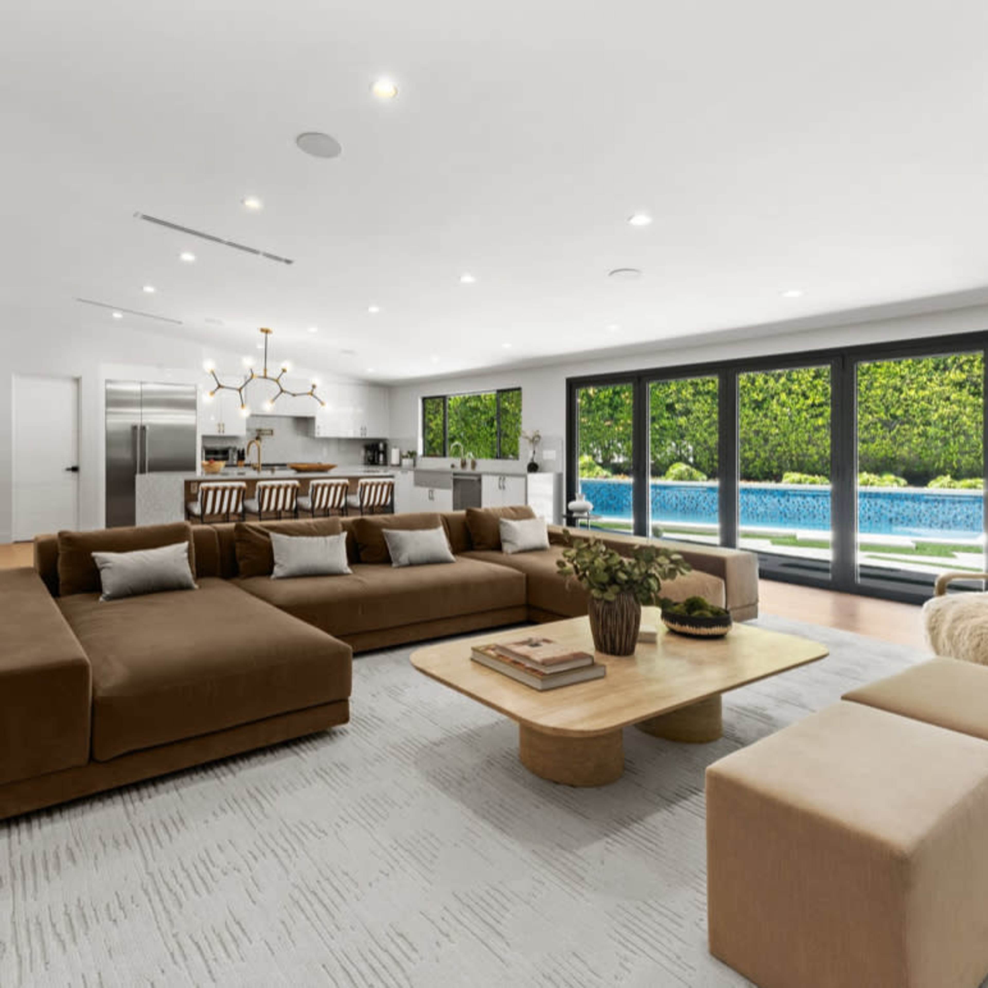 A modern open-concept living room with a large brown sectional sofa, a light coffee table, and a view of a swimming pool through expansive glass doors.