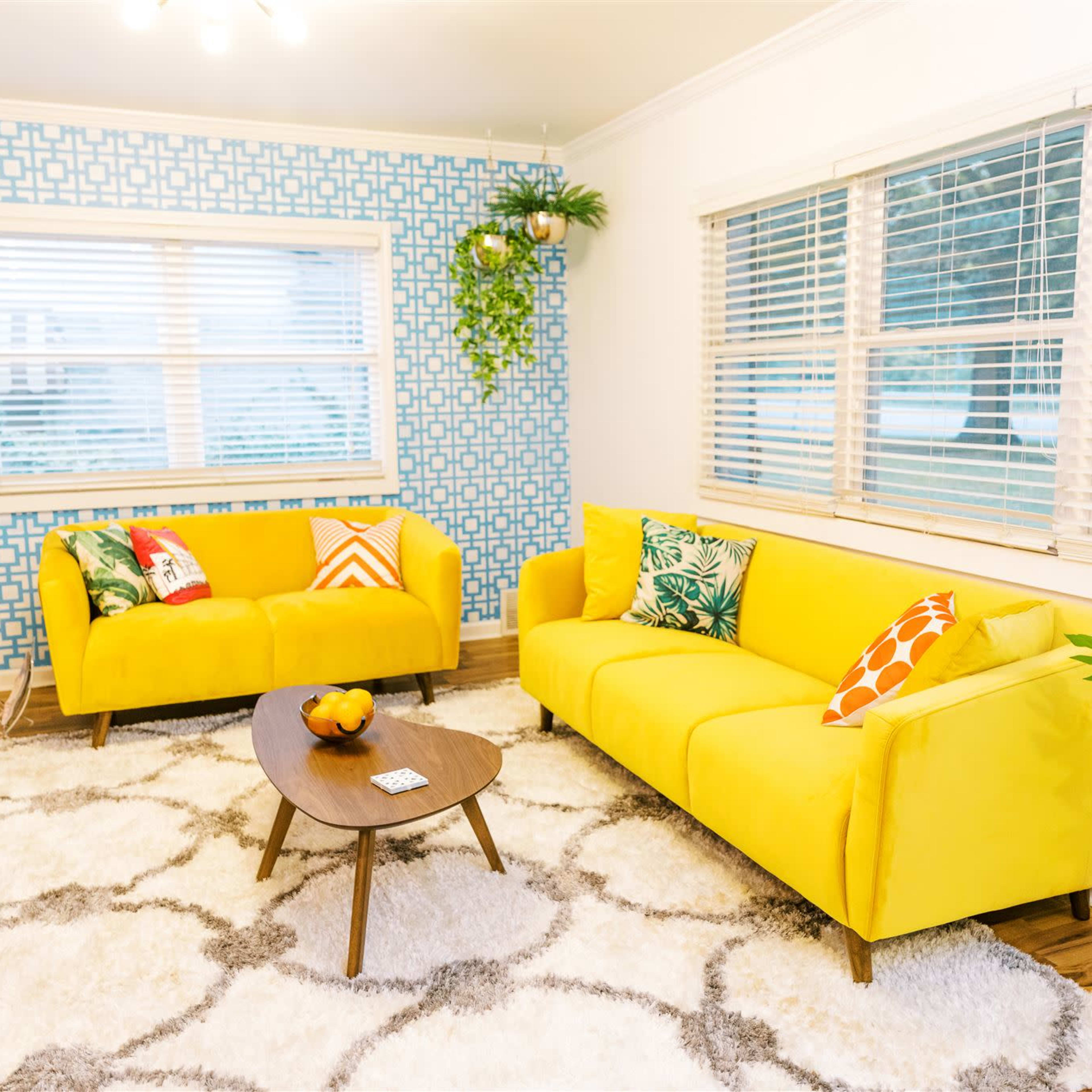 A brightly colored living room featuring yellow sofas, patterned cushions, a wooden coffee table, and a large area rug on a hardwood floor, with a patterned blue wall and large windows.