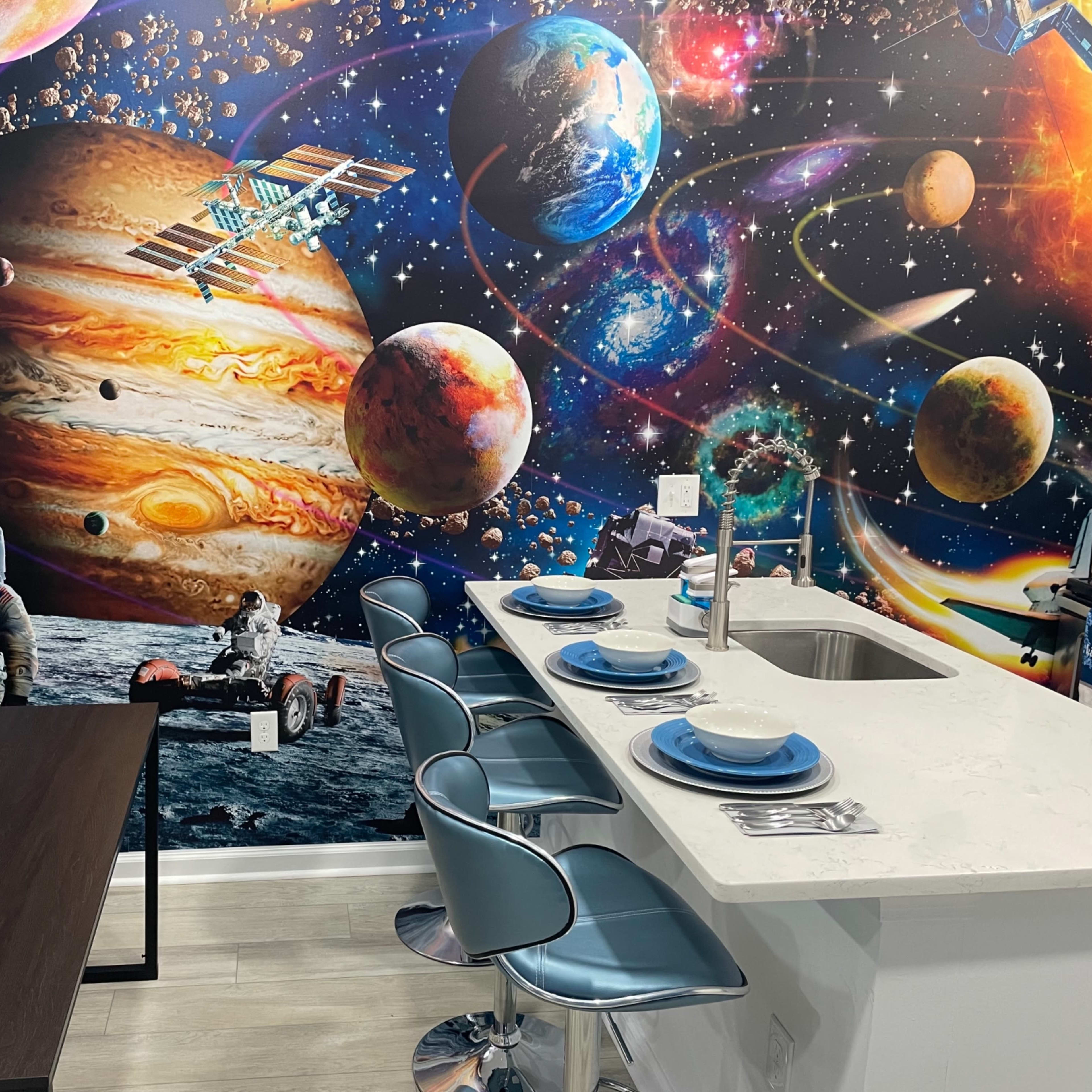 A modern kitchen with a white countertop and blue chairs is set against a vibrant space-themed mural featuring planets, stars, and a space shuttle.