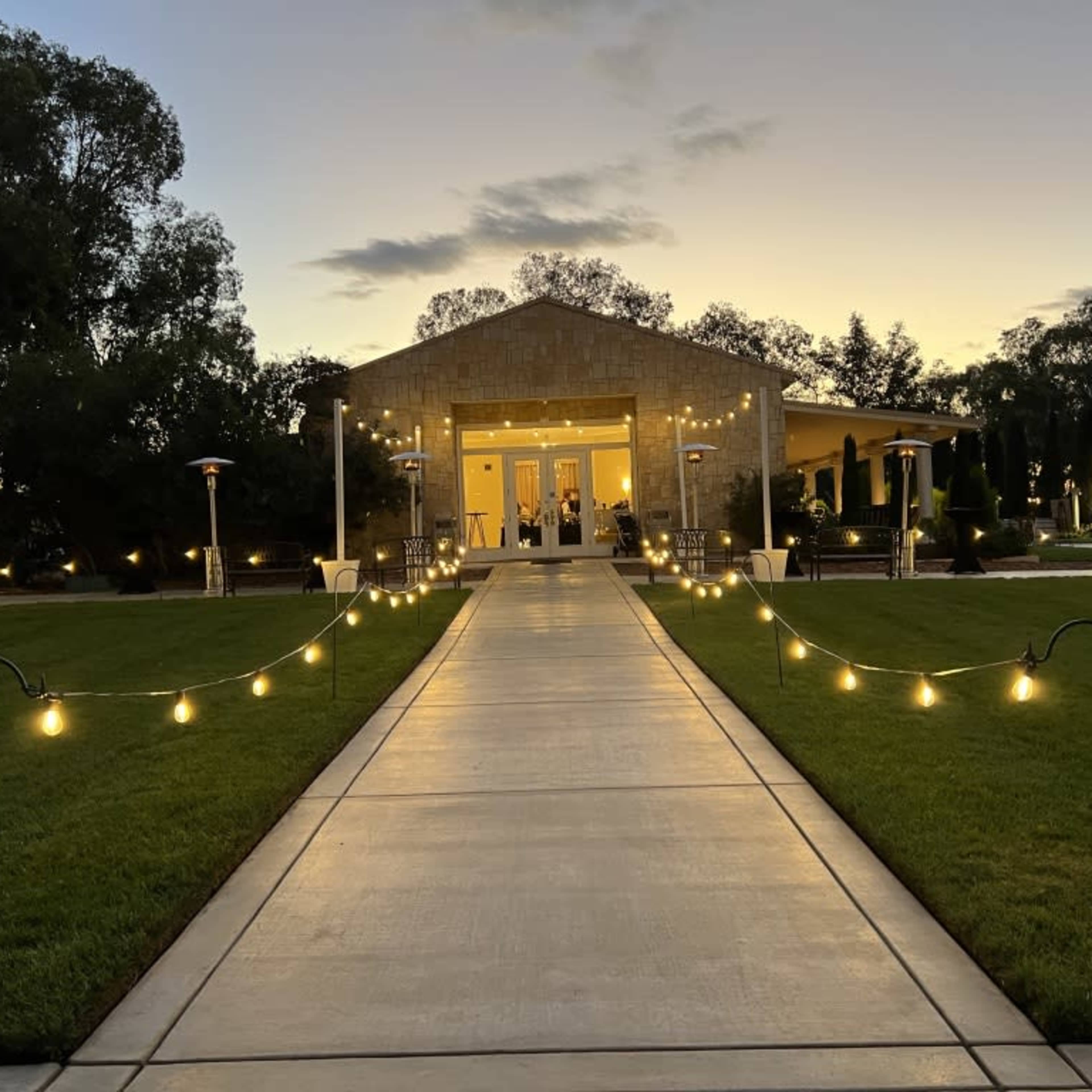 A pathway lined with lights leads to a large building at dusk, surrounded by neatly maintained greenery.