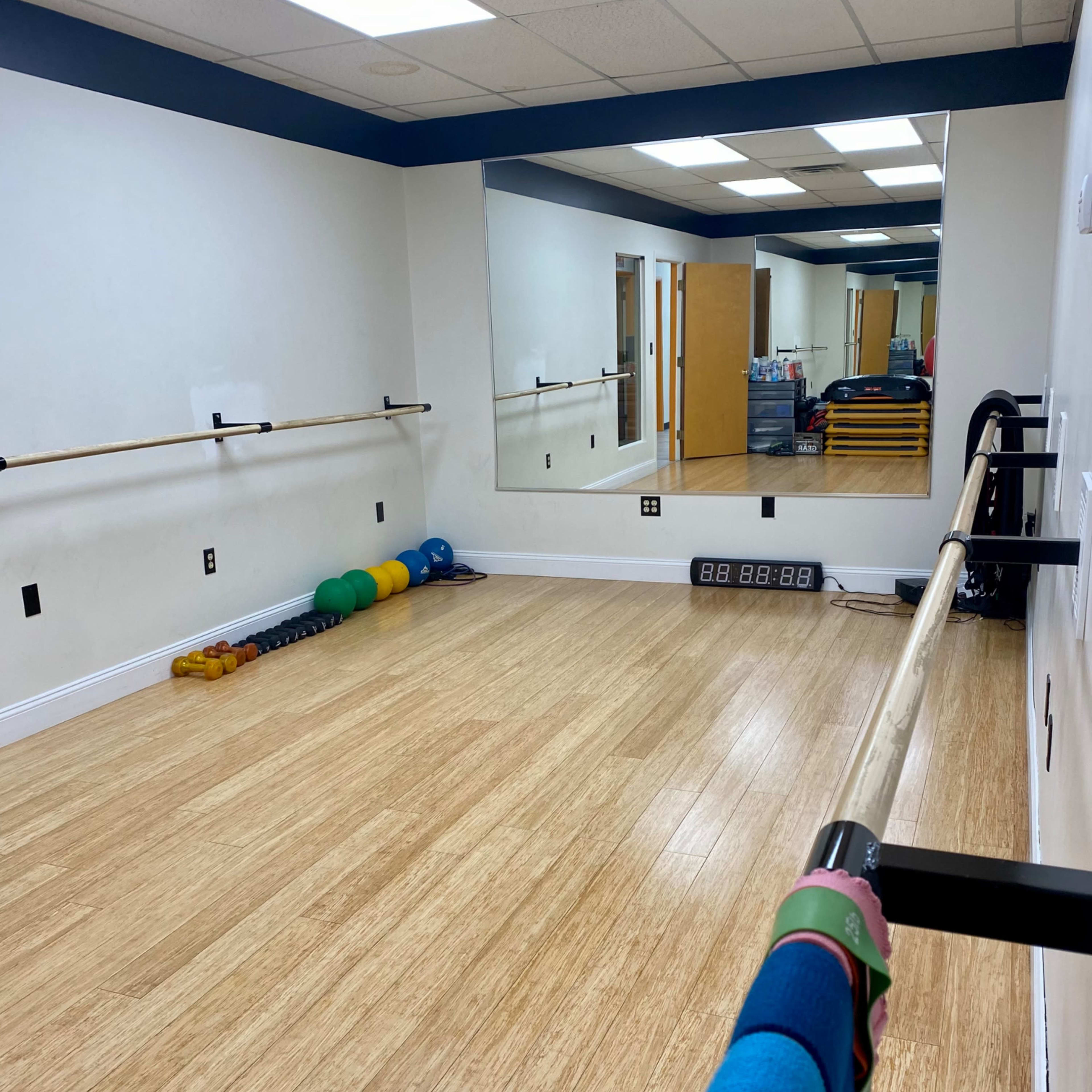 A fitness studio with wooden flooring, wall-mounted ballet barres, a large mirror, and exercise equipment arranged neatly along the wall.