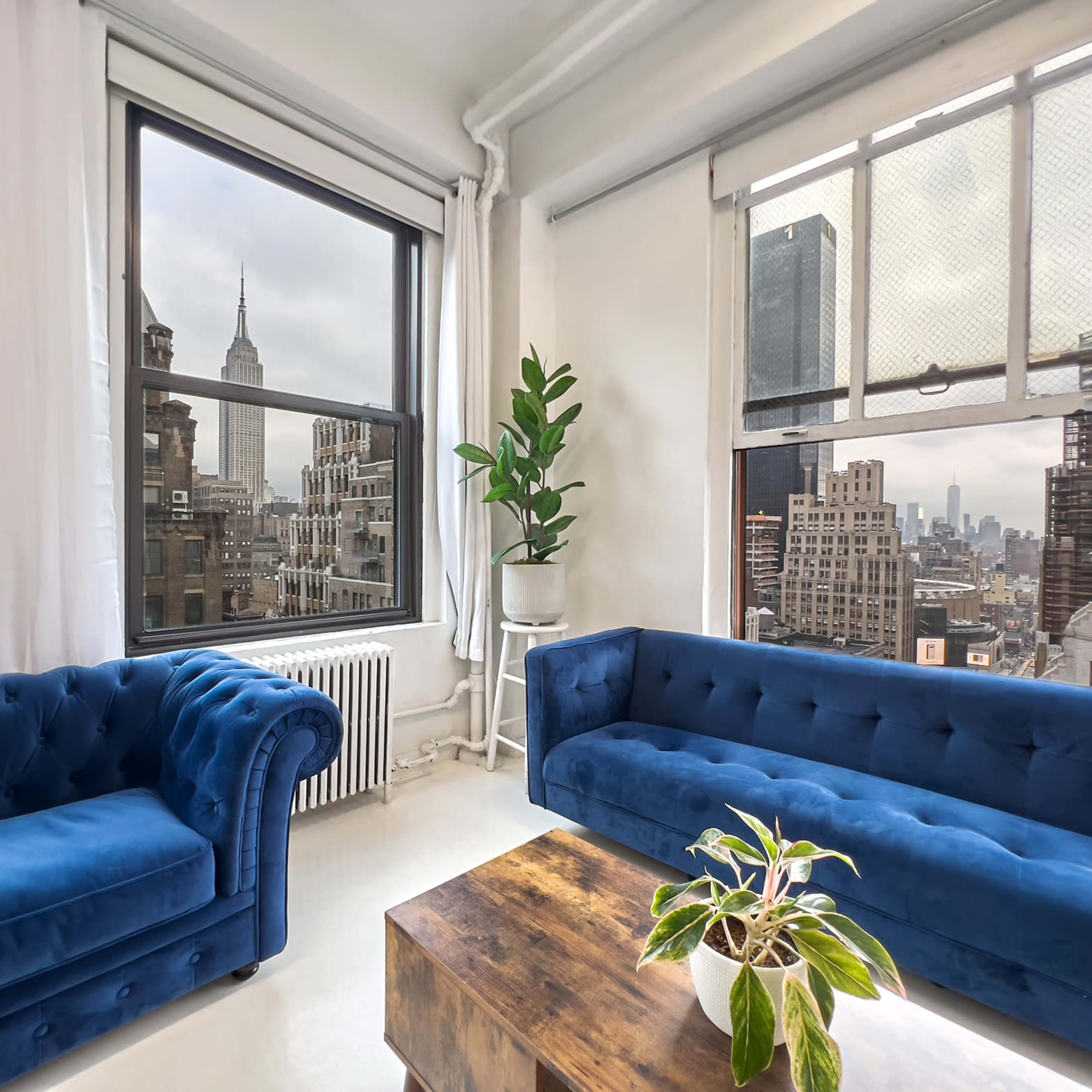 A cozy room with two blue velvet couches, a wooden coffee table, and a large window offering a view of the Empire State Building and city skyline.