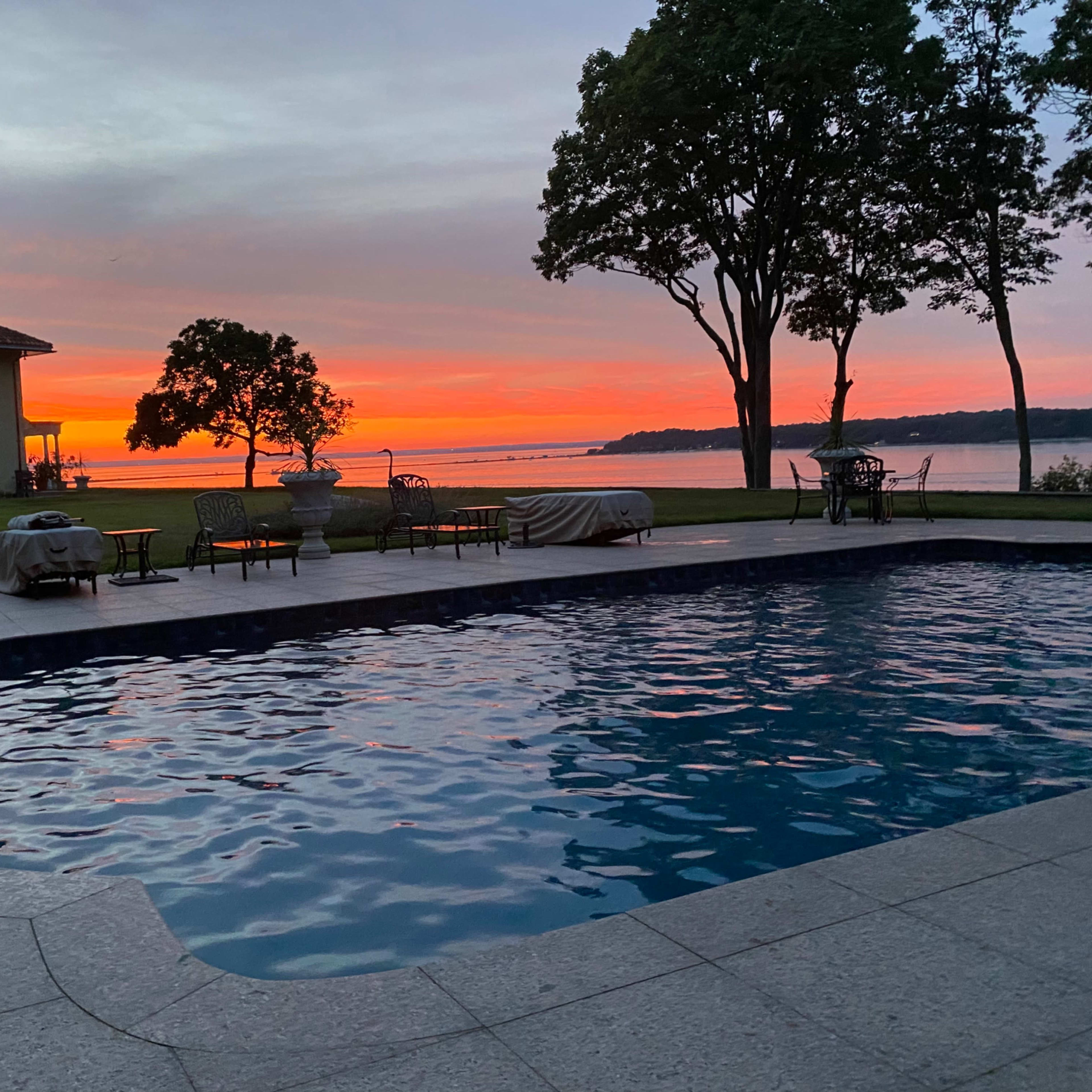 A swimming pool sits in the foreground with a view of a sunset sky over a body of water, framed by trees and a house in the background.