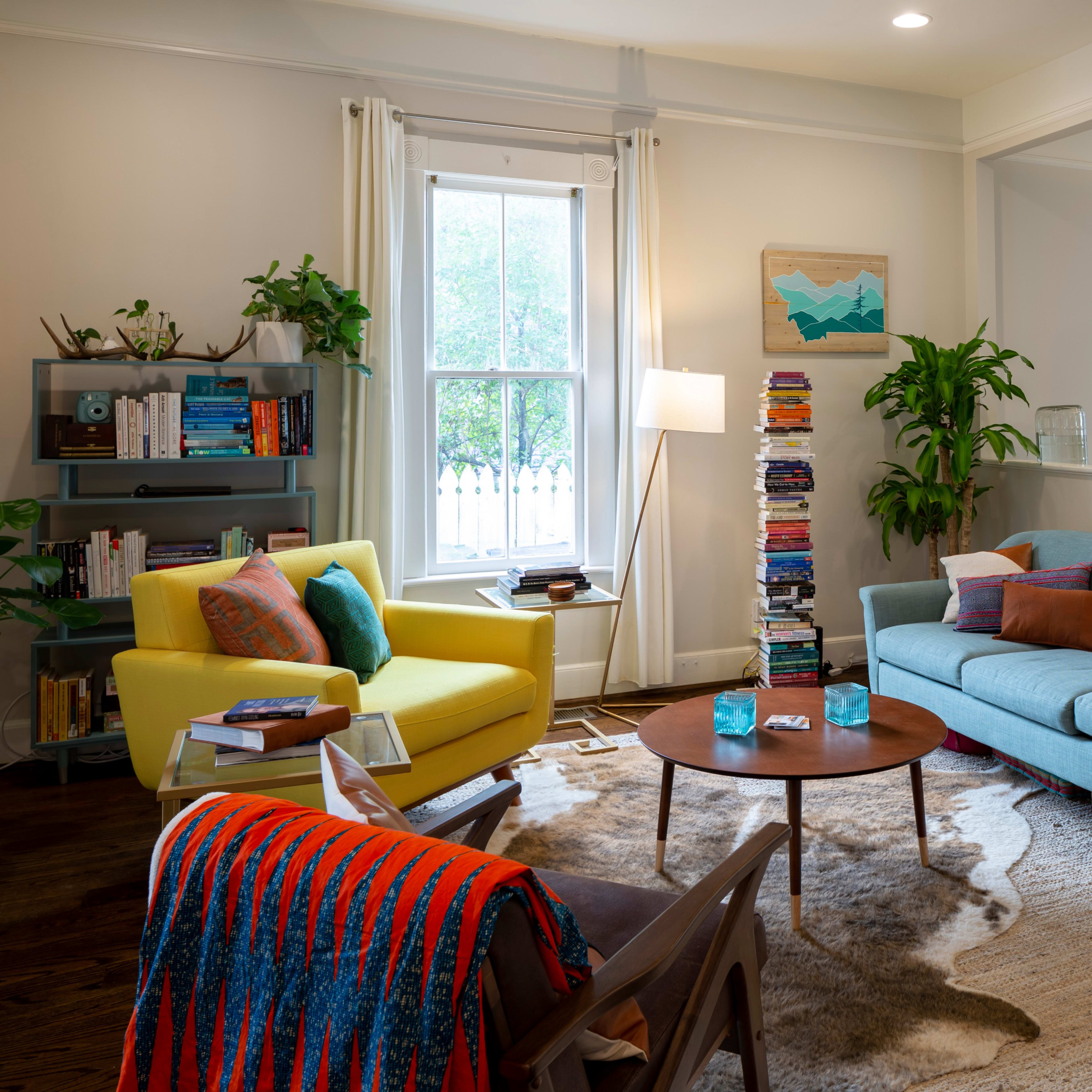 The living room features a blue sofa, a yellow armchair, a round coffee table, and a bookshelf filled with books, all arranged around a textured area rug.