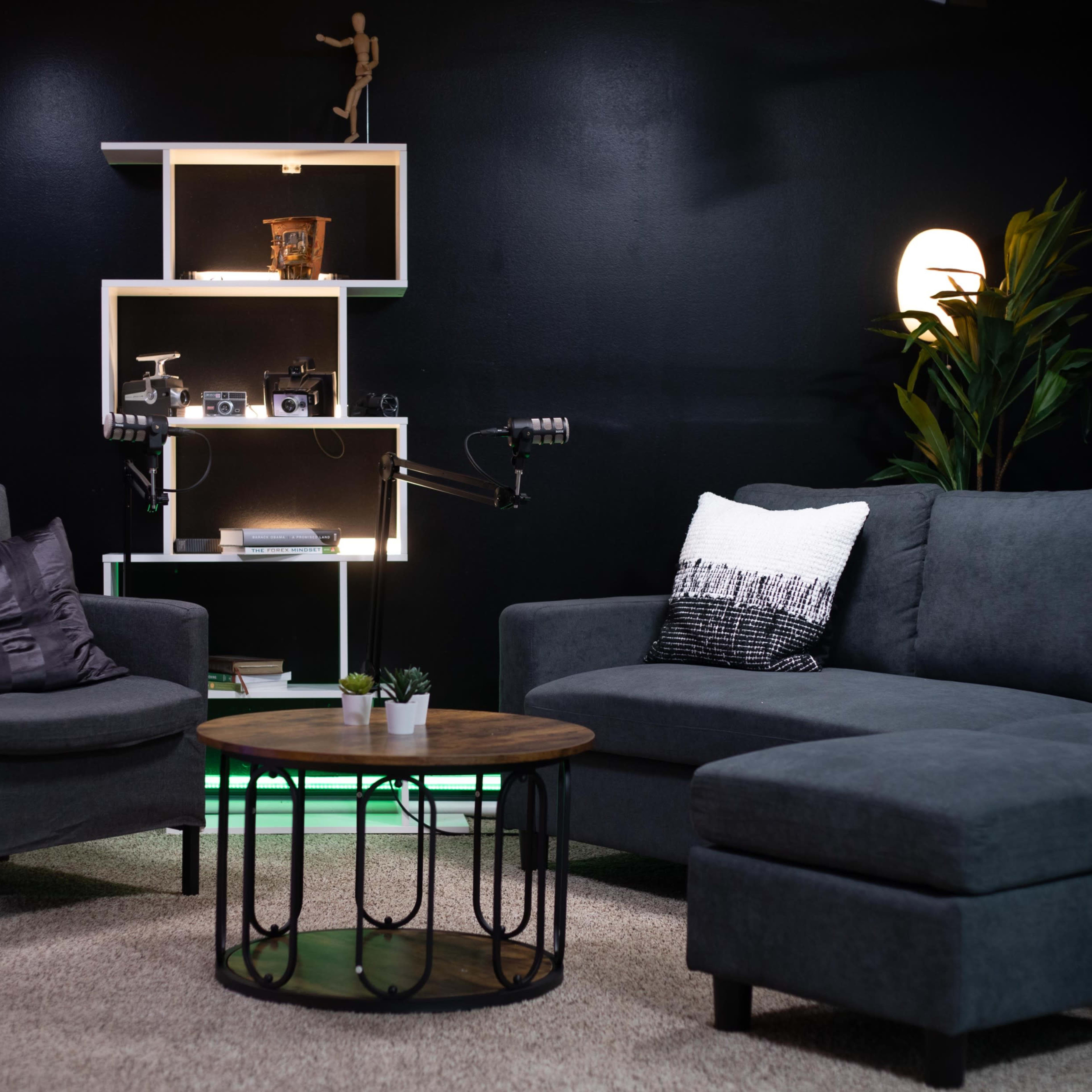 A modern living room setup featuring a dark gray sectional sofa, an armchair, a round wooden coffee table, and a shelf displaying decorative items against a black wall.