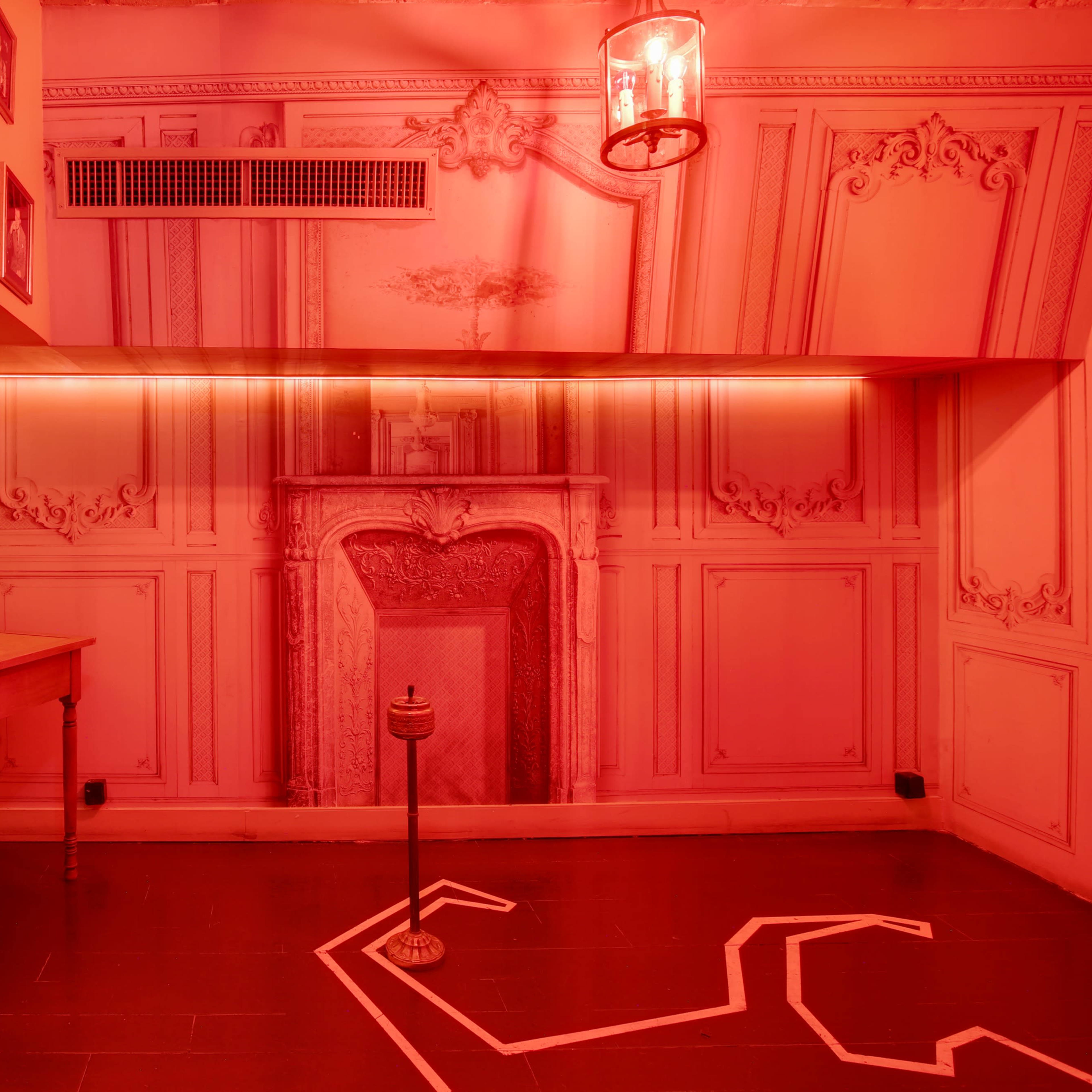 A small room with red lighting, featuring a wall mural of a fireplace, a wooden table, a chair, and a chalk outline on the floor.