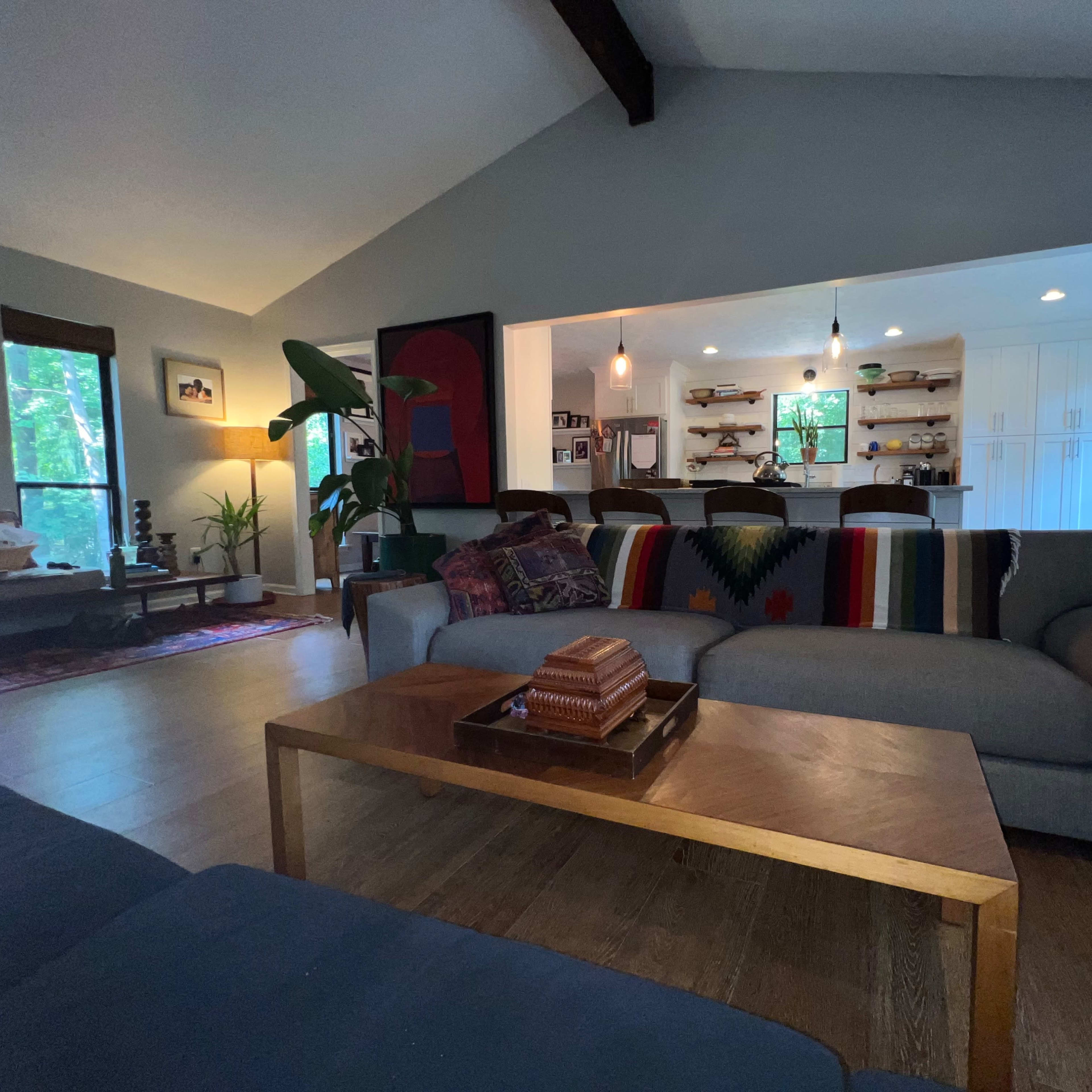 A living room with a sofa, a wooden coffee table, and an open kitchen in the background, featuring shelves and seating.