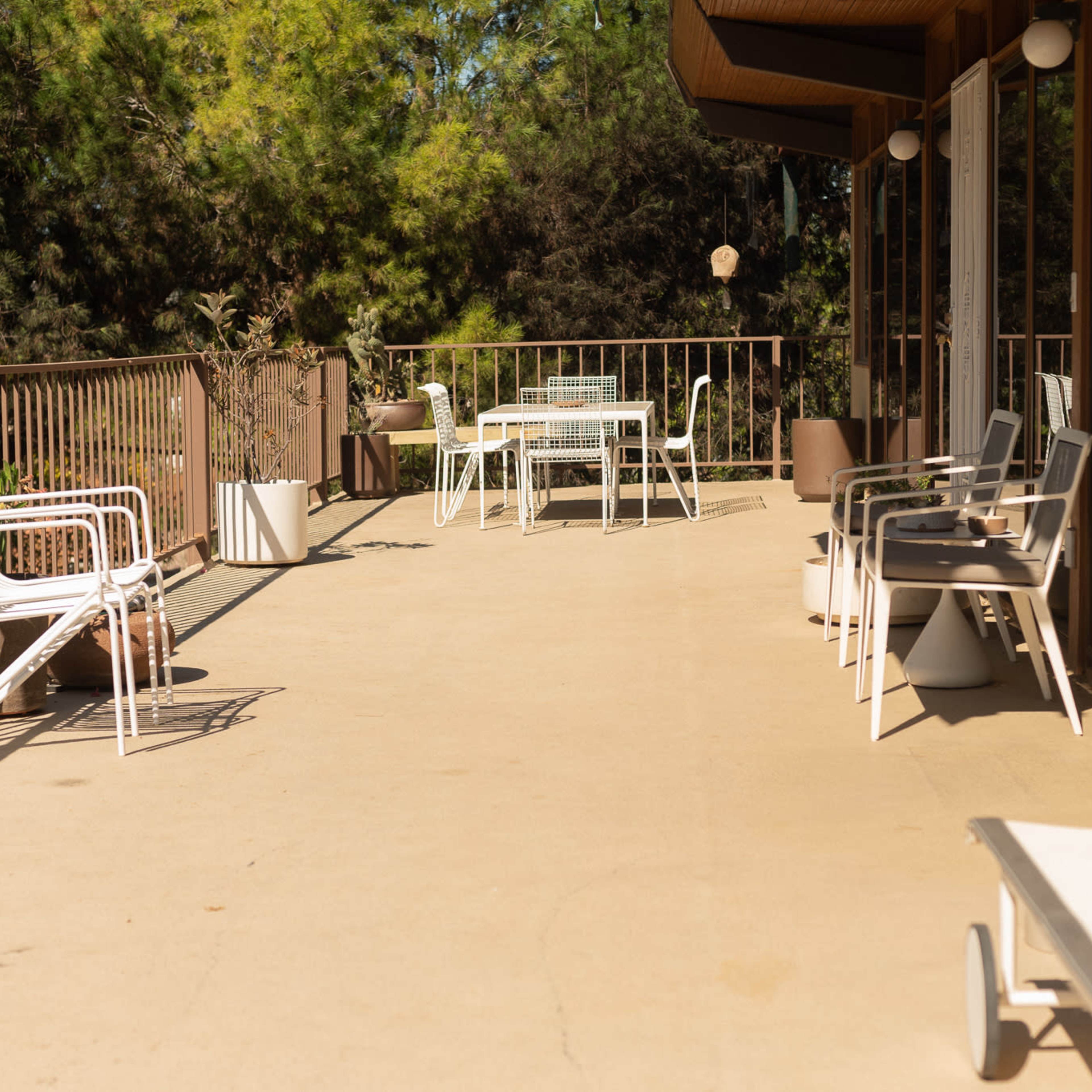 A sunlit patio with white outdoor furniture, including chairs and tables, set against a backdrop of greenery.