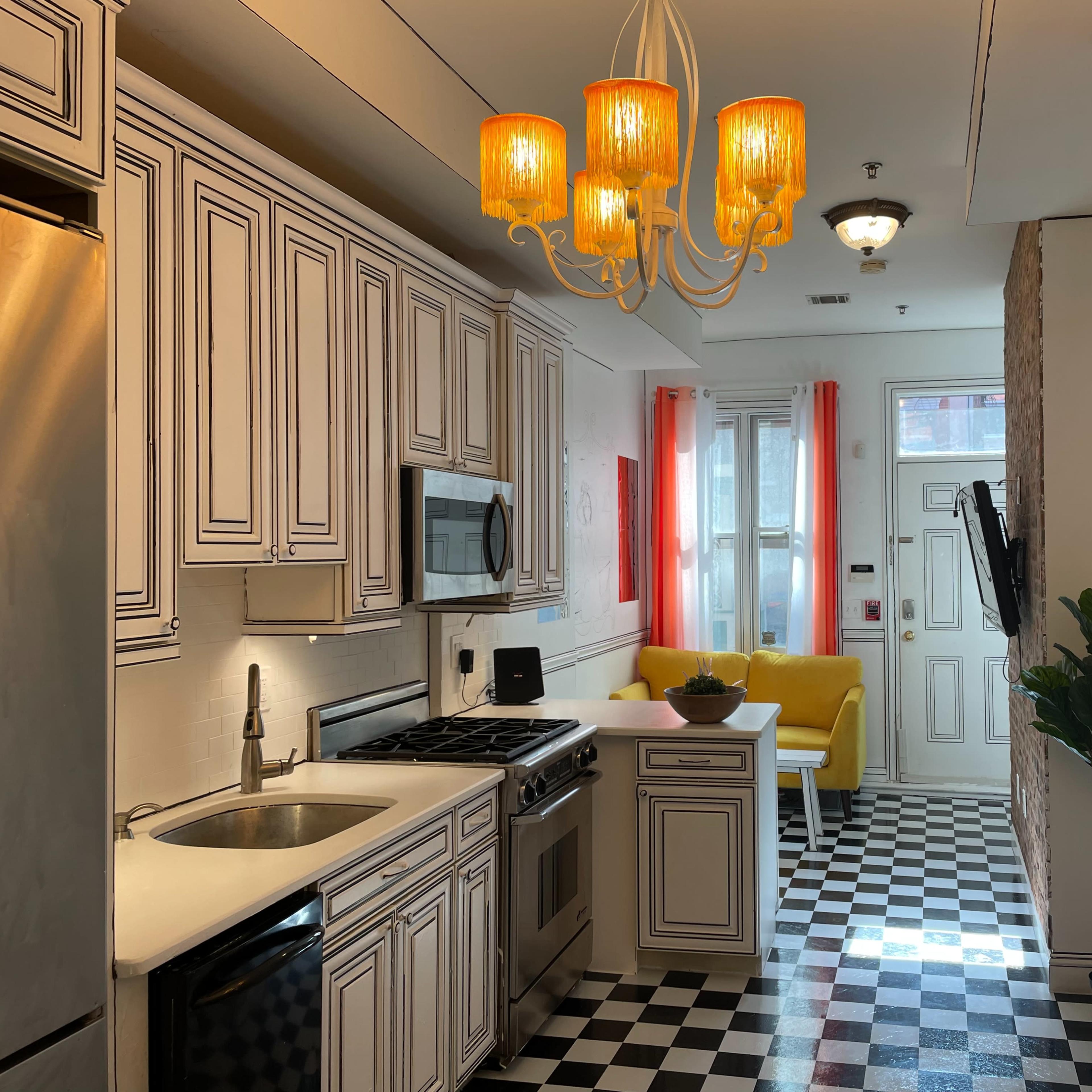 A kitchen with white cabinetry, a stainless steel refrigerator, and a checkered black-and-white floor, leading into a bright living area with a yellow sofa.