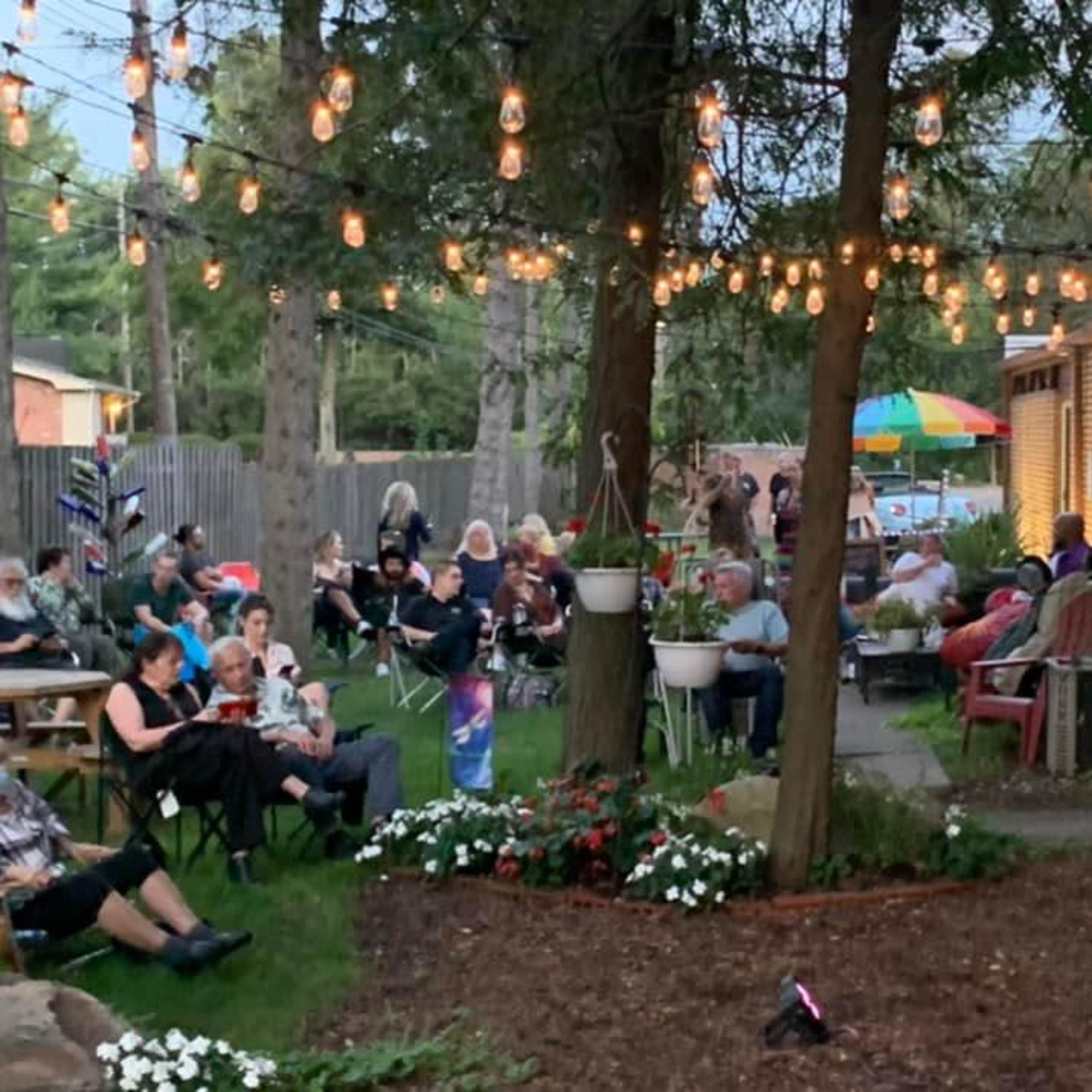 A gathering of people sits on chairs and benches in a yard, surrounded by trees and string lights, with a patio and colorful umbrellas in the background.