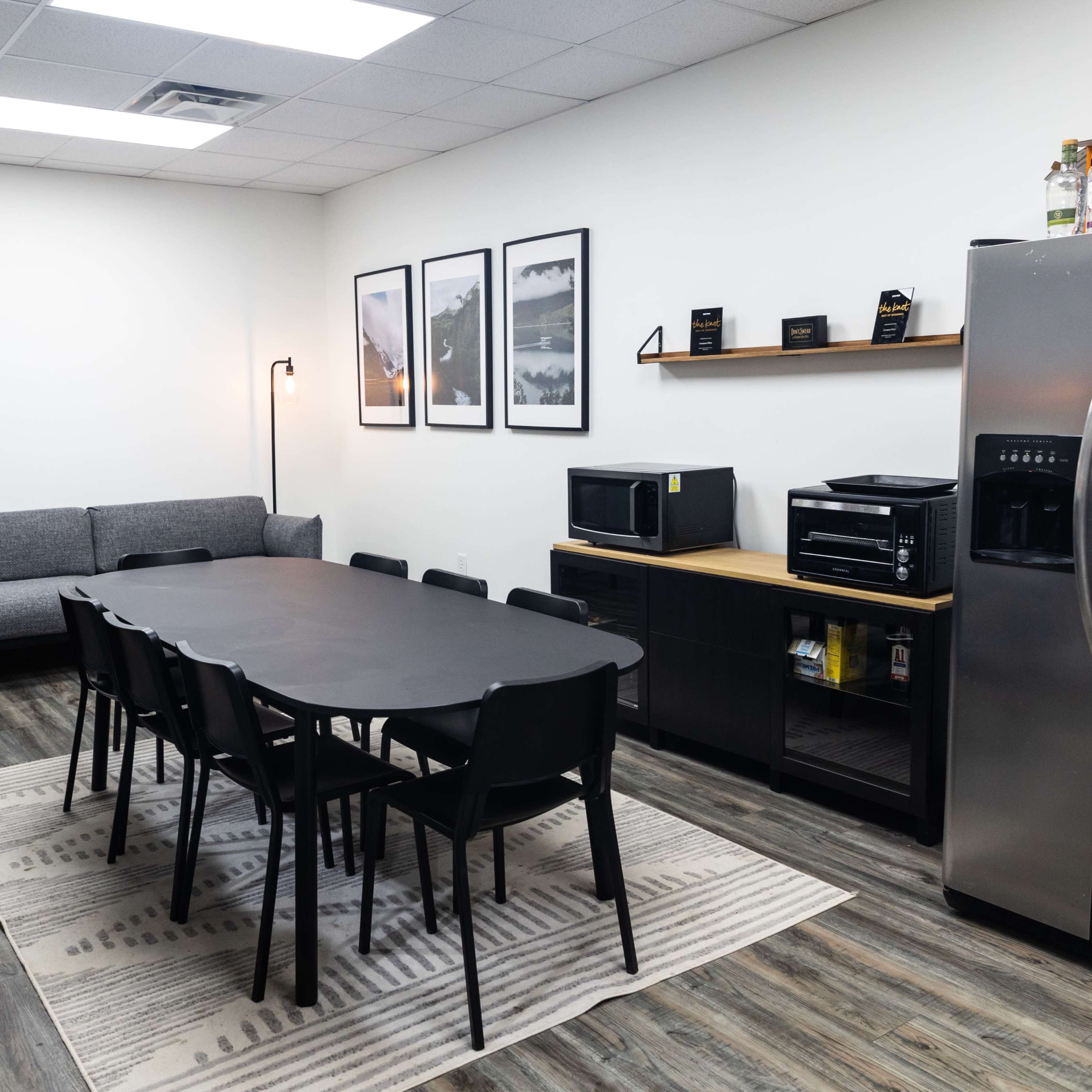 A modern break room with a long black table surrounded by chairs, a couch, kitchen appliances, and framed artwork on the walls.