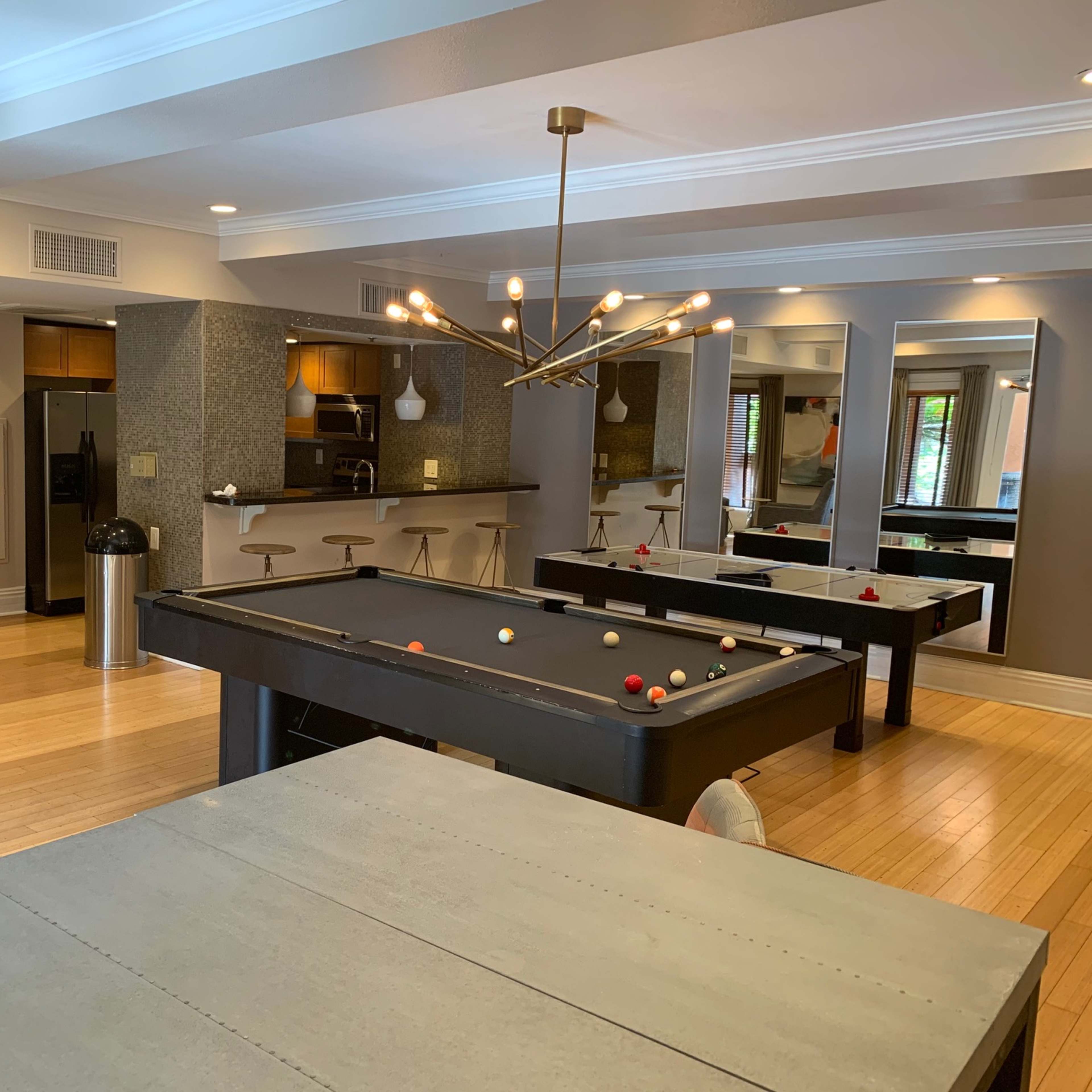 A modern recreational room with a pool table, dining tables, and a kitchen area, featuring hardwood floors and contemporary lighting.