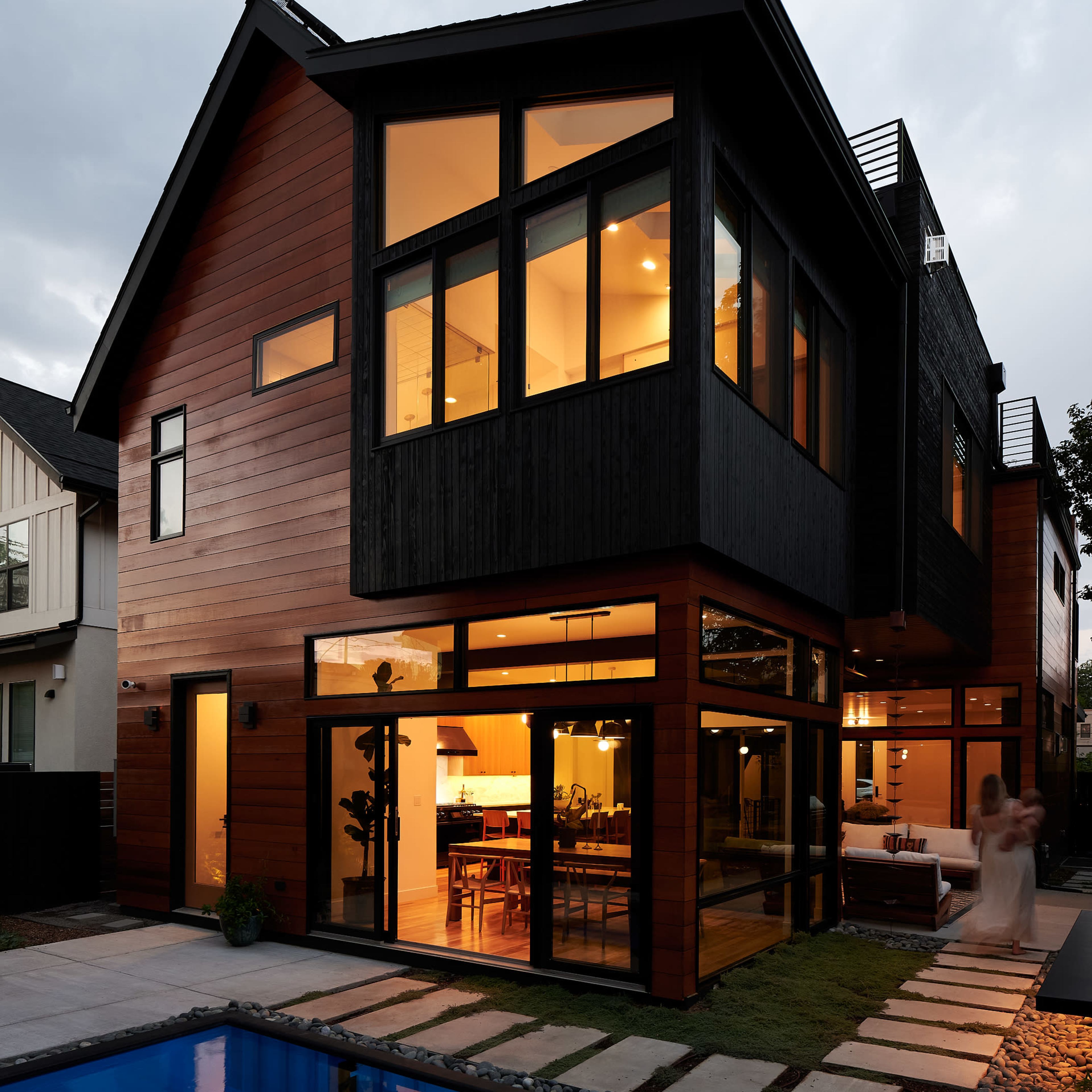 A modern two-story house with large windows overlooking a blue swimming pool and a pathway lined with stepping stones.