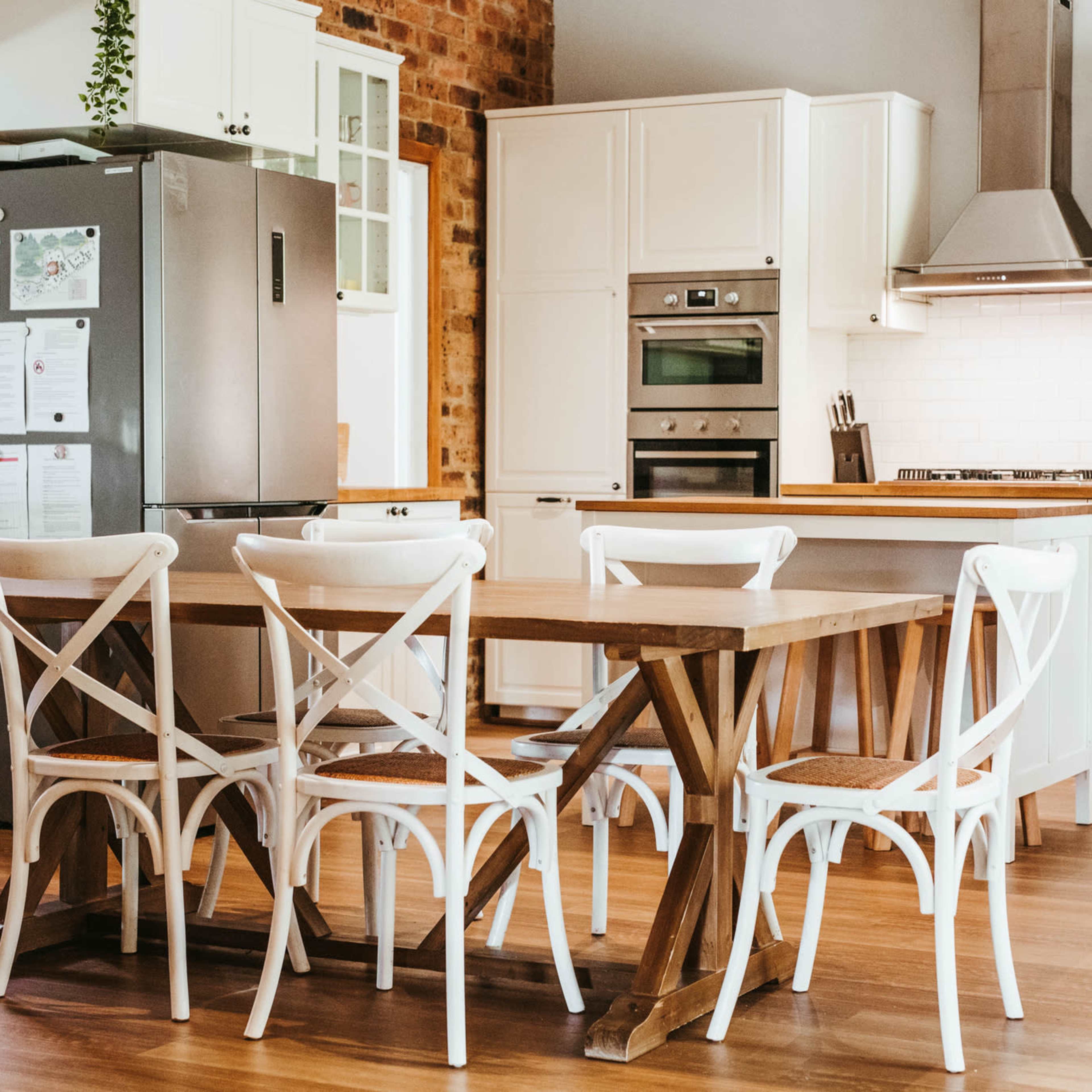 A spacious kitchen features a wooden dining table surrounded by white chairs, with a modern refrigerator and cooking appliances in the background.
