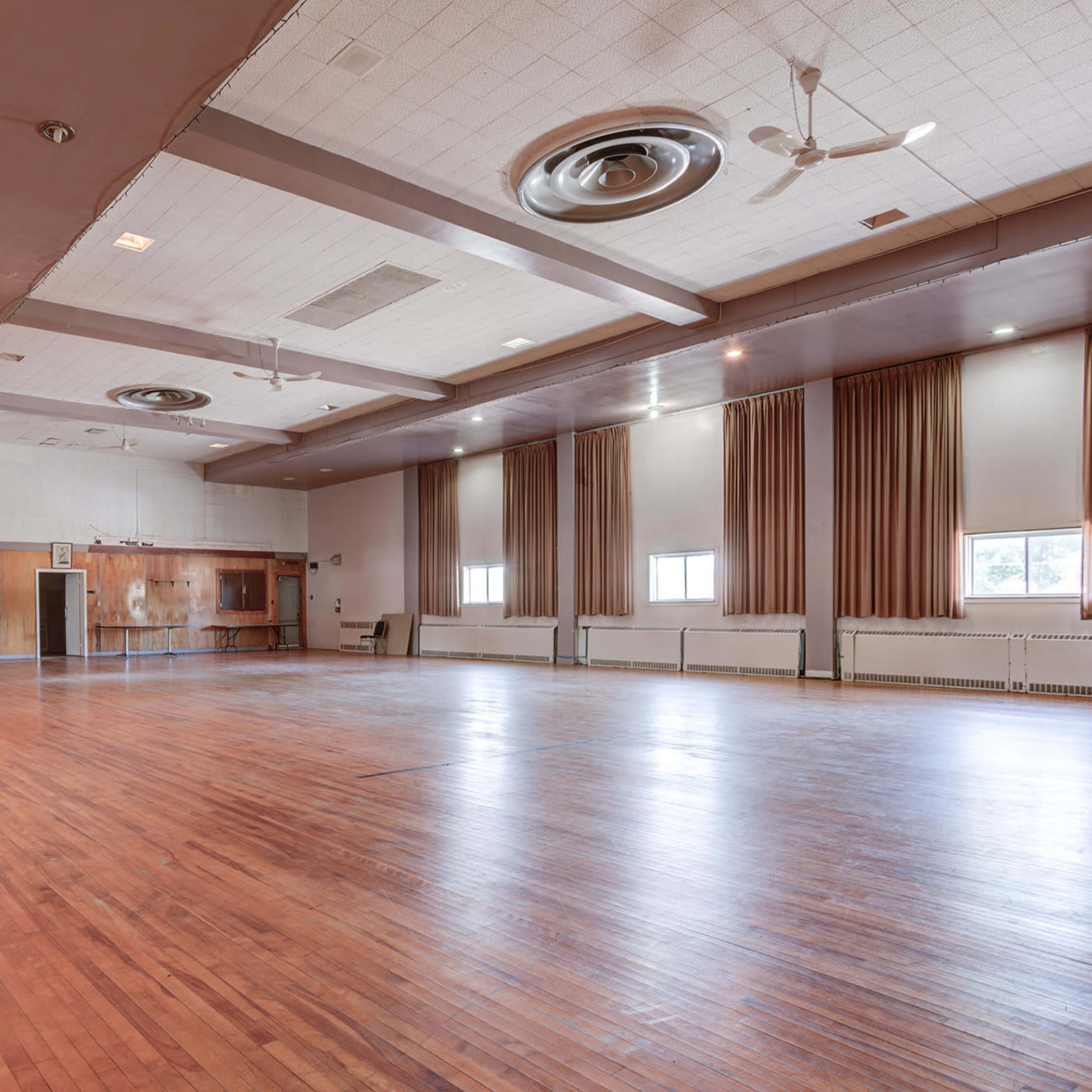 A spacious, empty hall with a polished wooden floor, large windows, and ceiling fans.