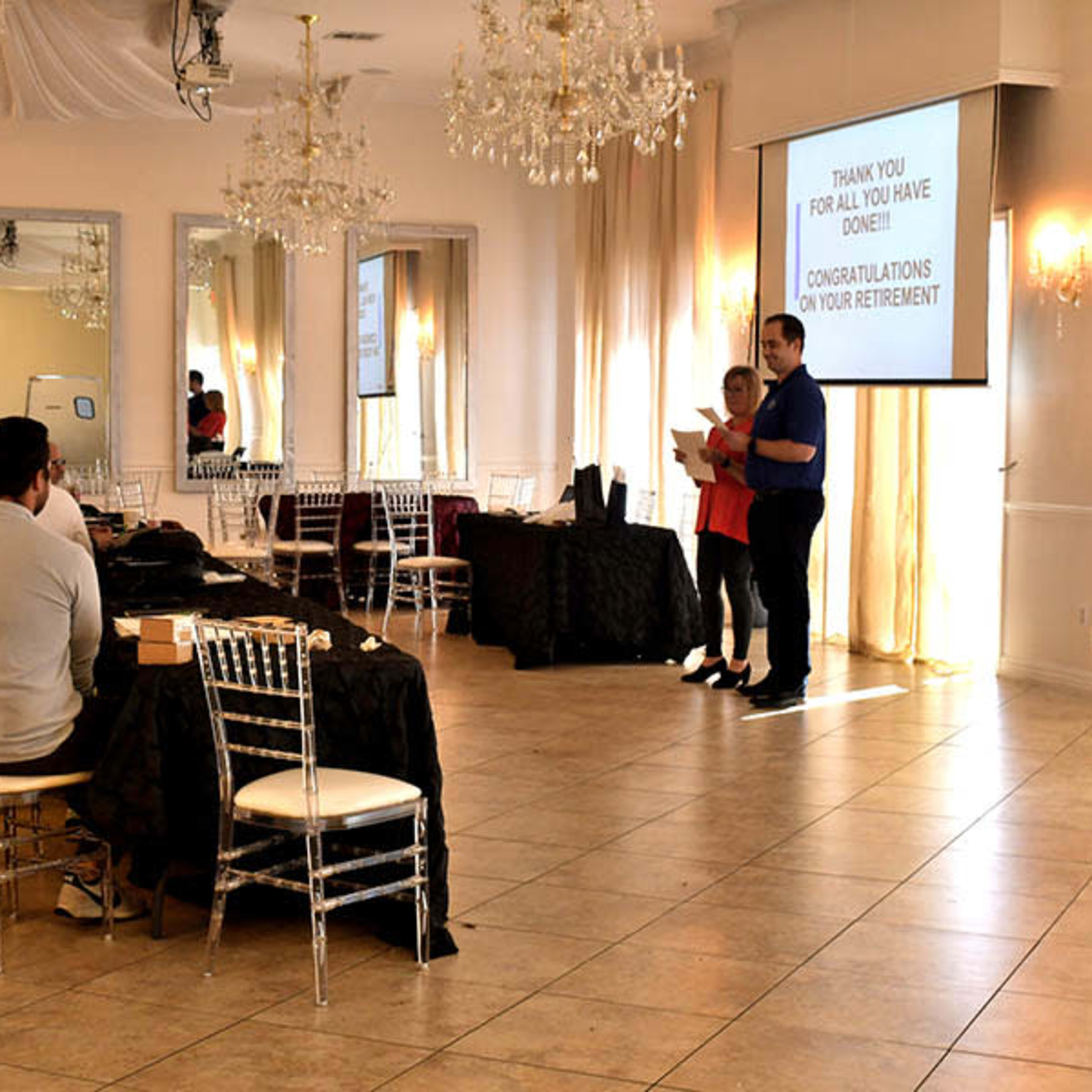 A man and a woman stand in front of a screen displaying a retirement message in a decorated hall with tables and chairs arranged for an event.