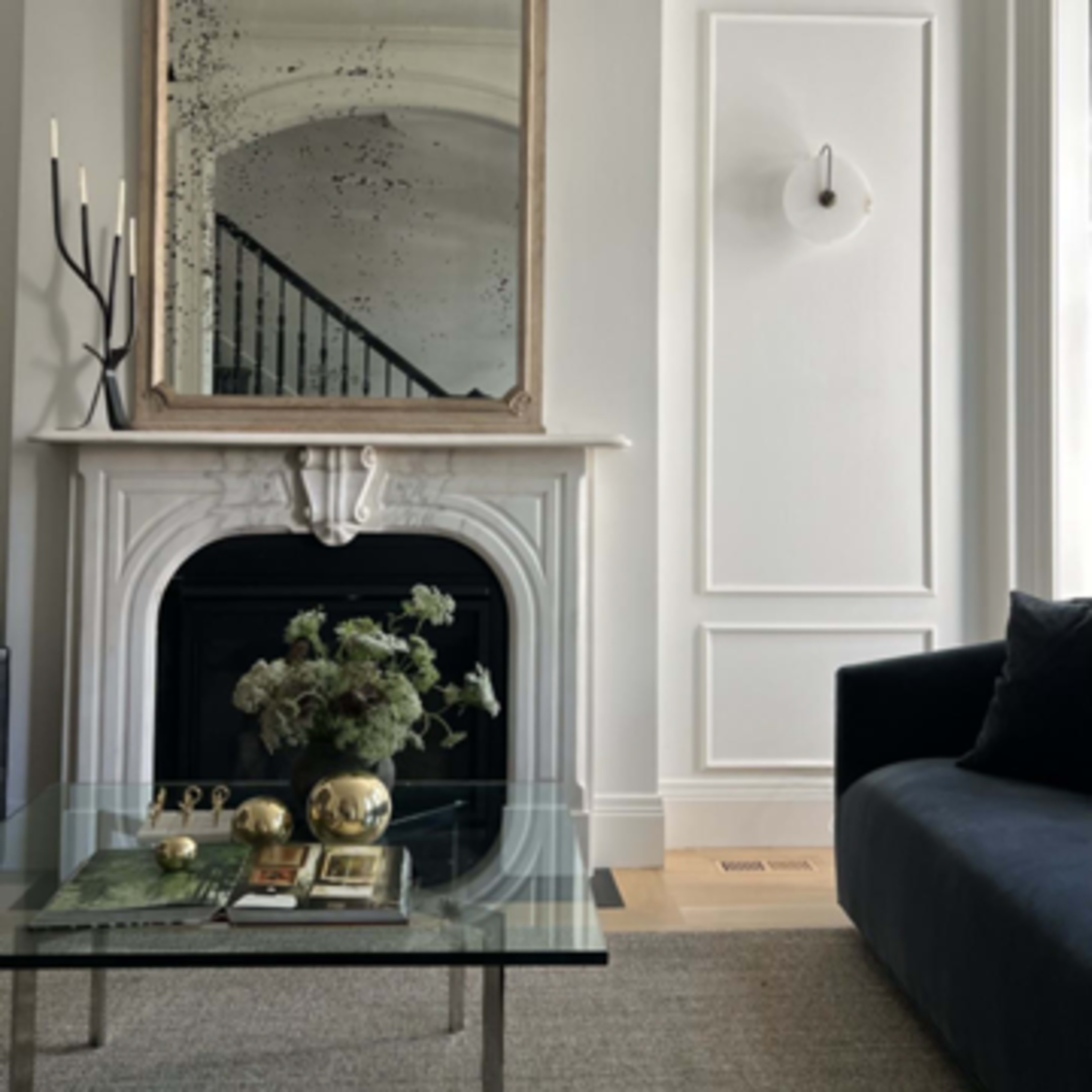 A modern living room featuring a glass coffee table in front of a black sofa, a decorative mirror above a white mantel, and a staircase visible in the background.