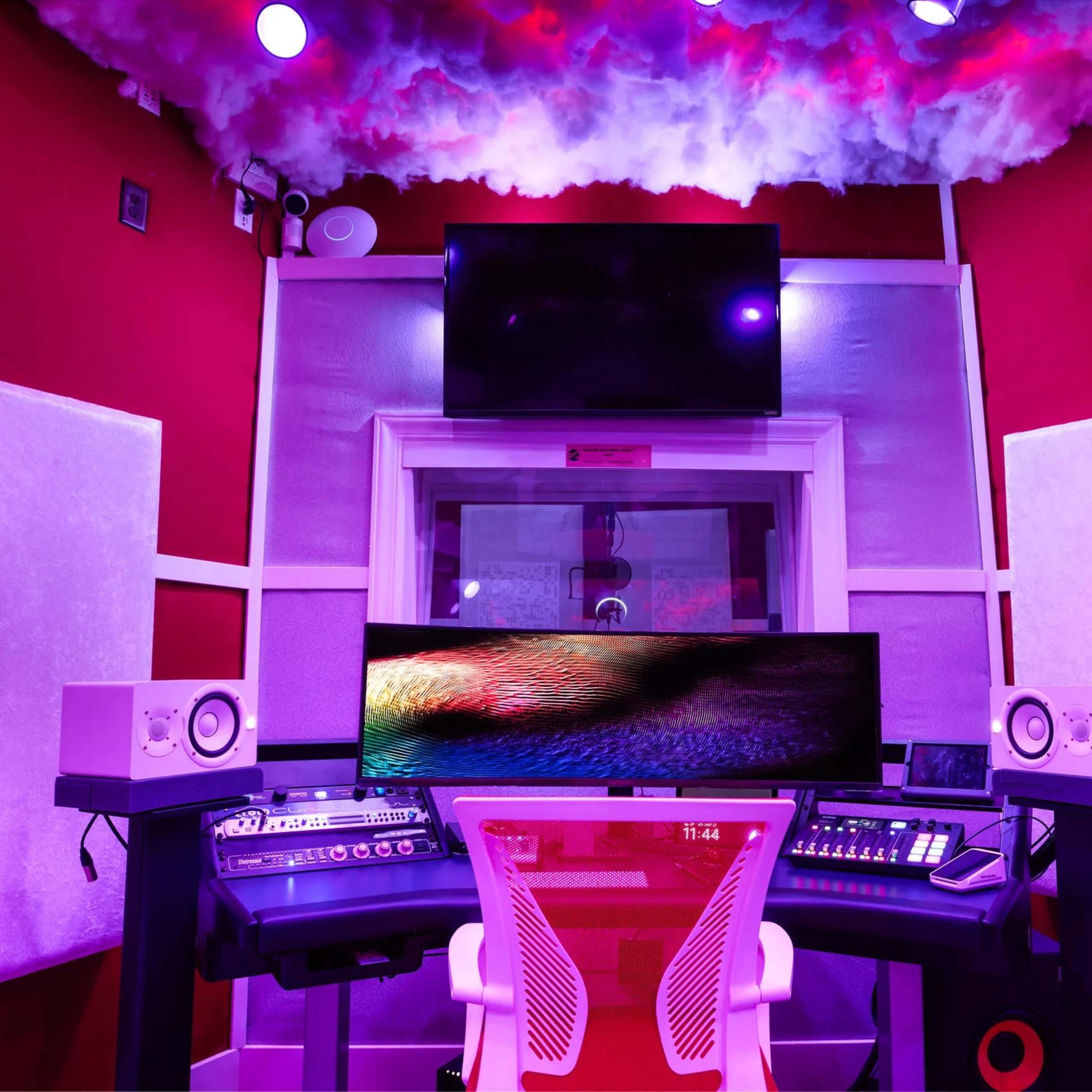 A music studio features a central desk with audio equipment, a large monitor, and soundproofing panels against bright red walls.